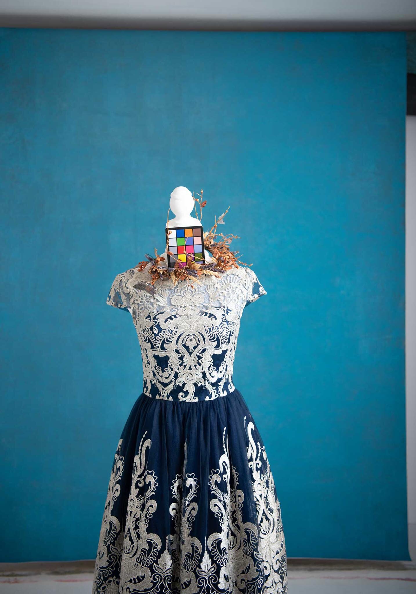 An art installation features a dress on a dress form with a color calibration chart on the chest. The dress has a navy and white intricate floral pattern, and the background is a plain blue wall.