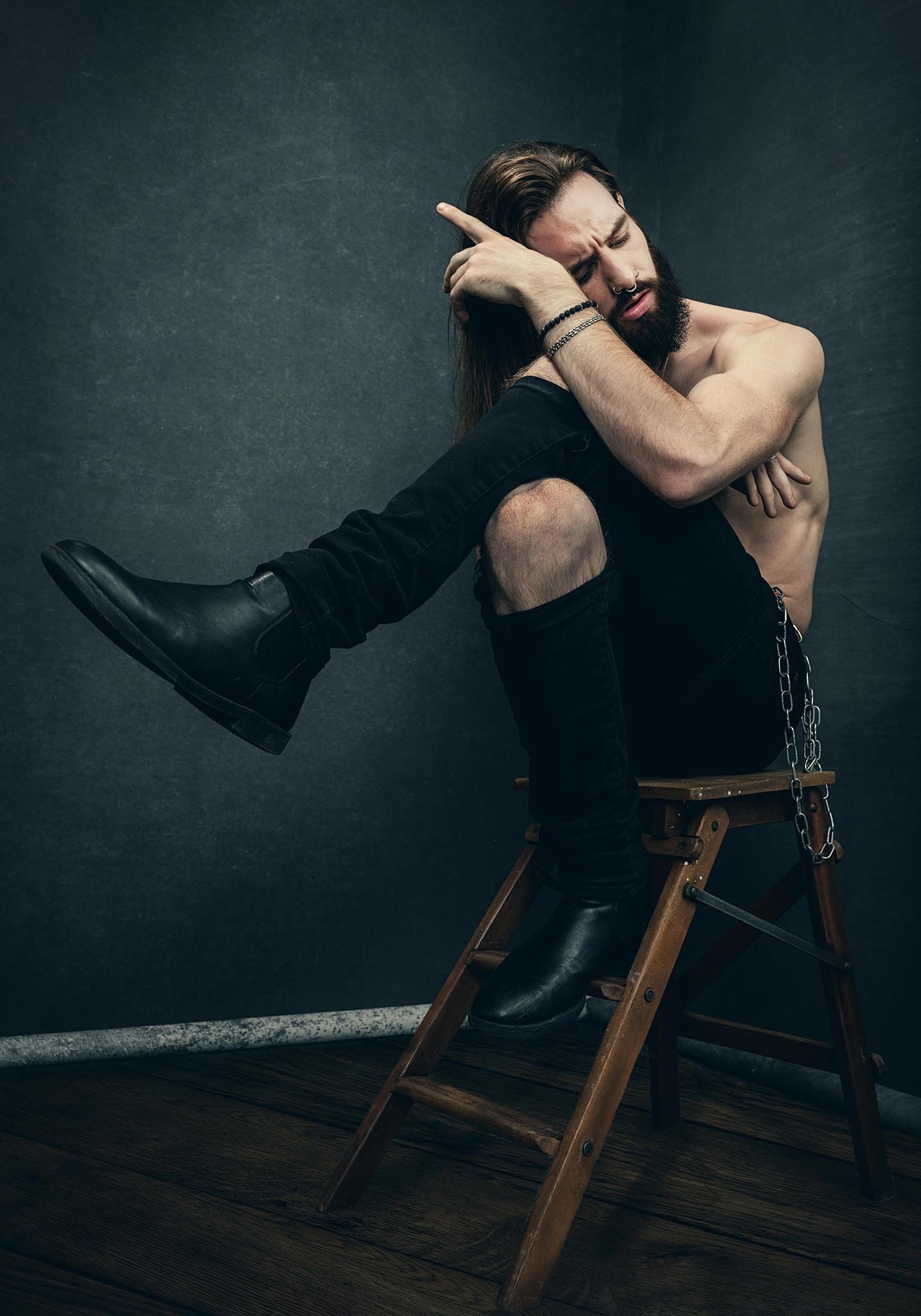 A shirtless man with long hair and a beard sitting on a wooden chair, with one leg resting on the chair and knee pulled up, in a dark room with a plain wall, exhibiting a thoughtful or pensive expression.