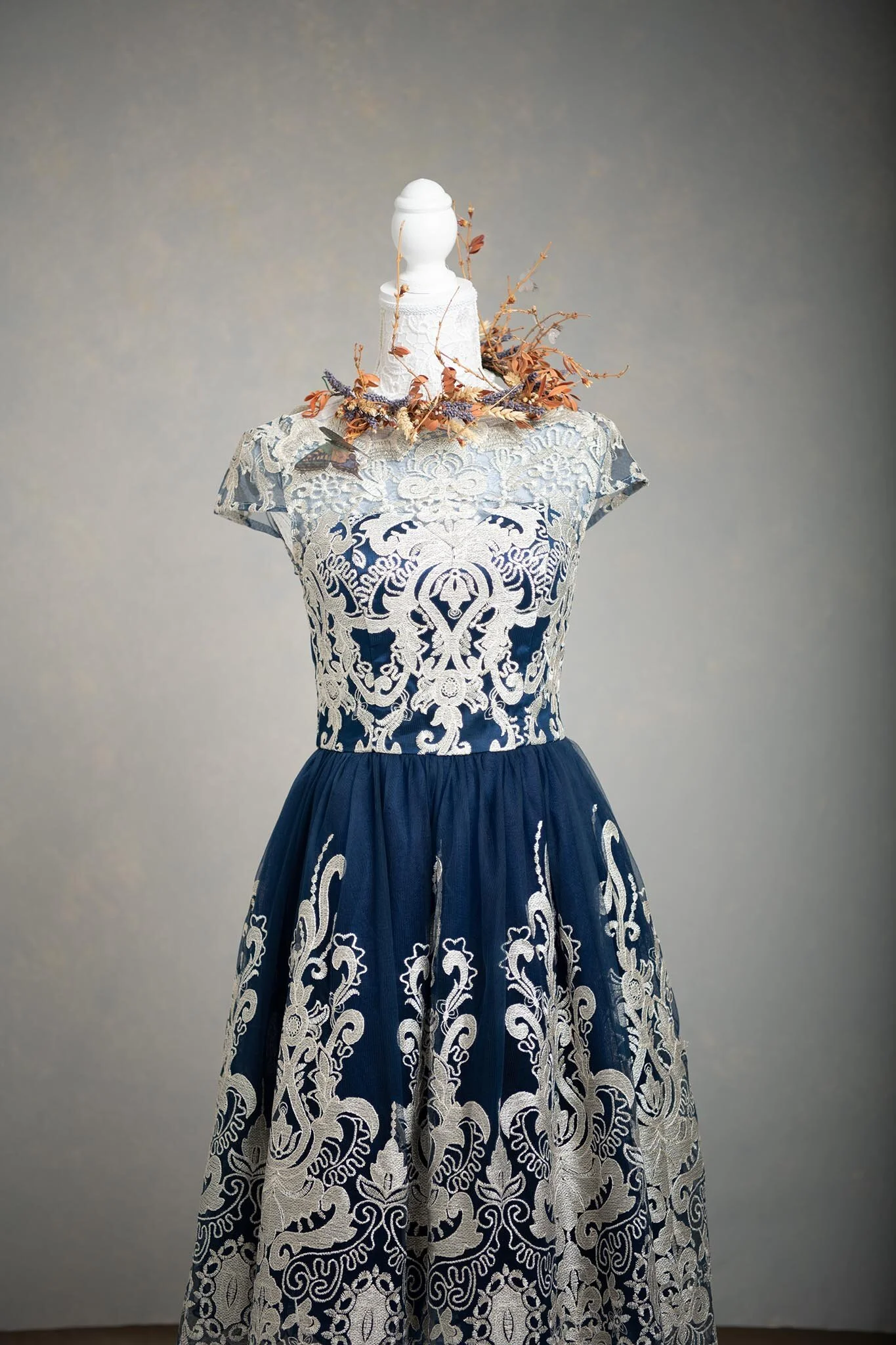 A dress on a mannequin featuring ornate white lace embroidery and navy blue fabric with intricate floral and scroll patterns.