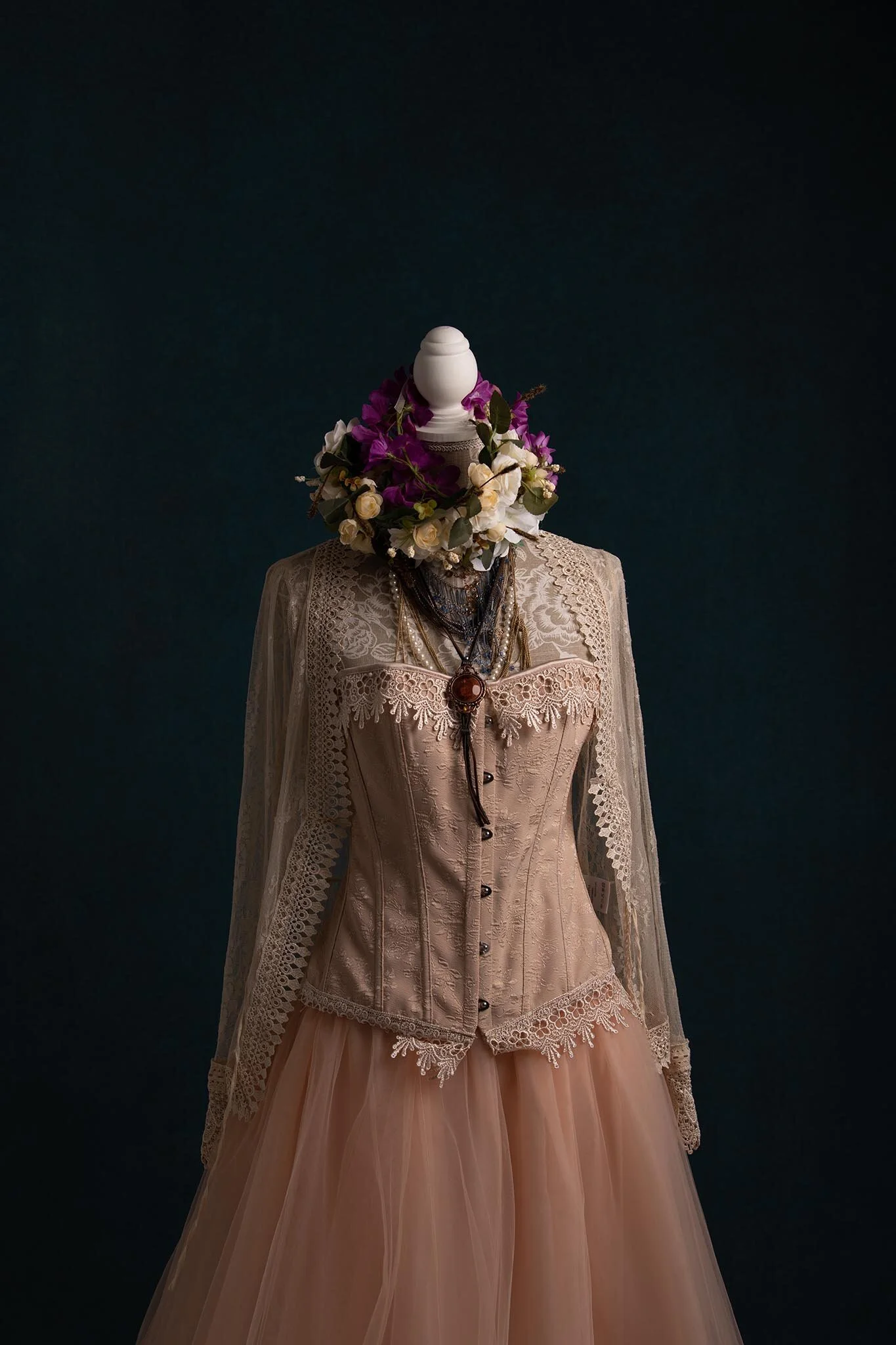A mannequin dressed in vintage bridal attire with a lacy, beige top and pink tulle skirt, adorned with a floral headpiece made of white and purple flowers, and layered necklaces against a dark background.