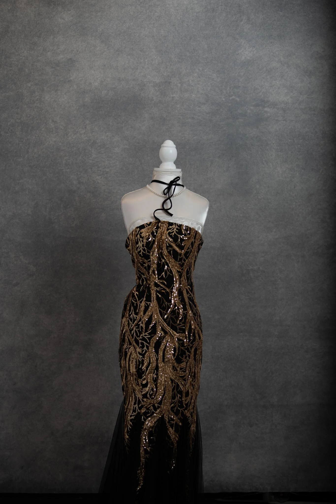 A mannequin wearing a strapless, black and gold sequined gown with intricate gold embroidery, displayed against a gray textured background.