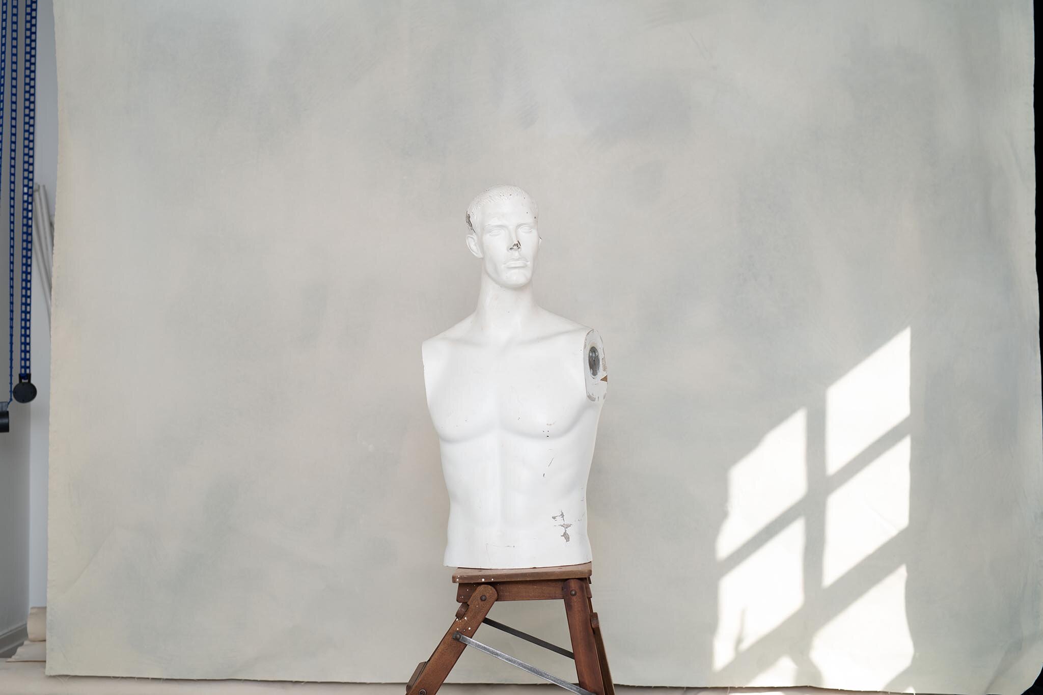 White mannequin torso on a wooden easel with shadowed window light on background wall.