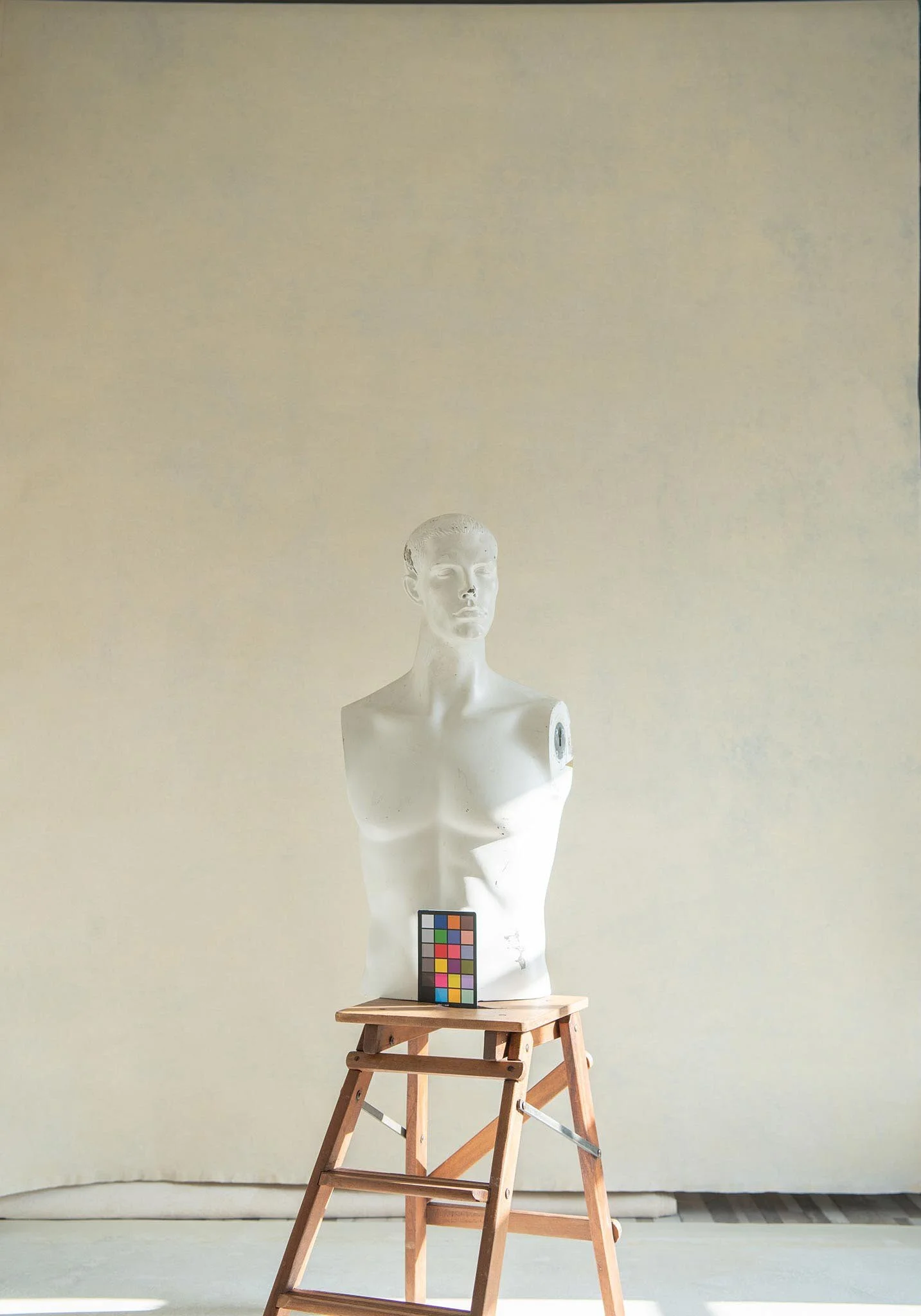 A white female mannequin bust on a wooden stepladder, with a color calibration card in front, set against a plain, light-colored background.