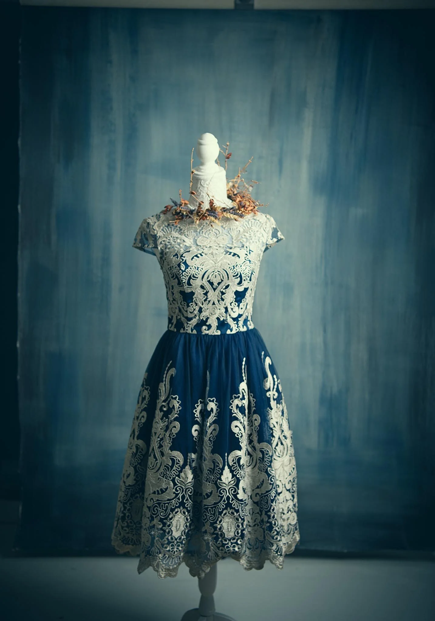 A vintage dress on a mannequin with intricate lace and embroidery, displayed in front of a textured blue wall.