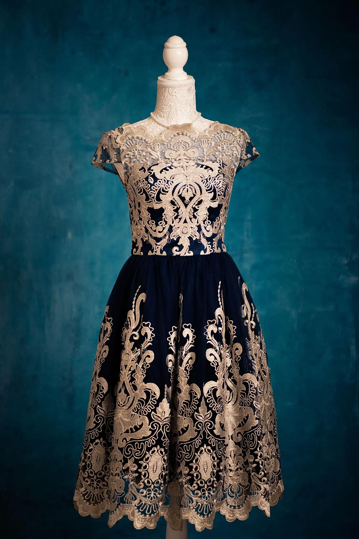 A vintage dress with intricate cream lace overlay on a dark blue fabric, displayed on a headless mannequin against a textured blue background.