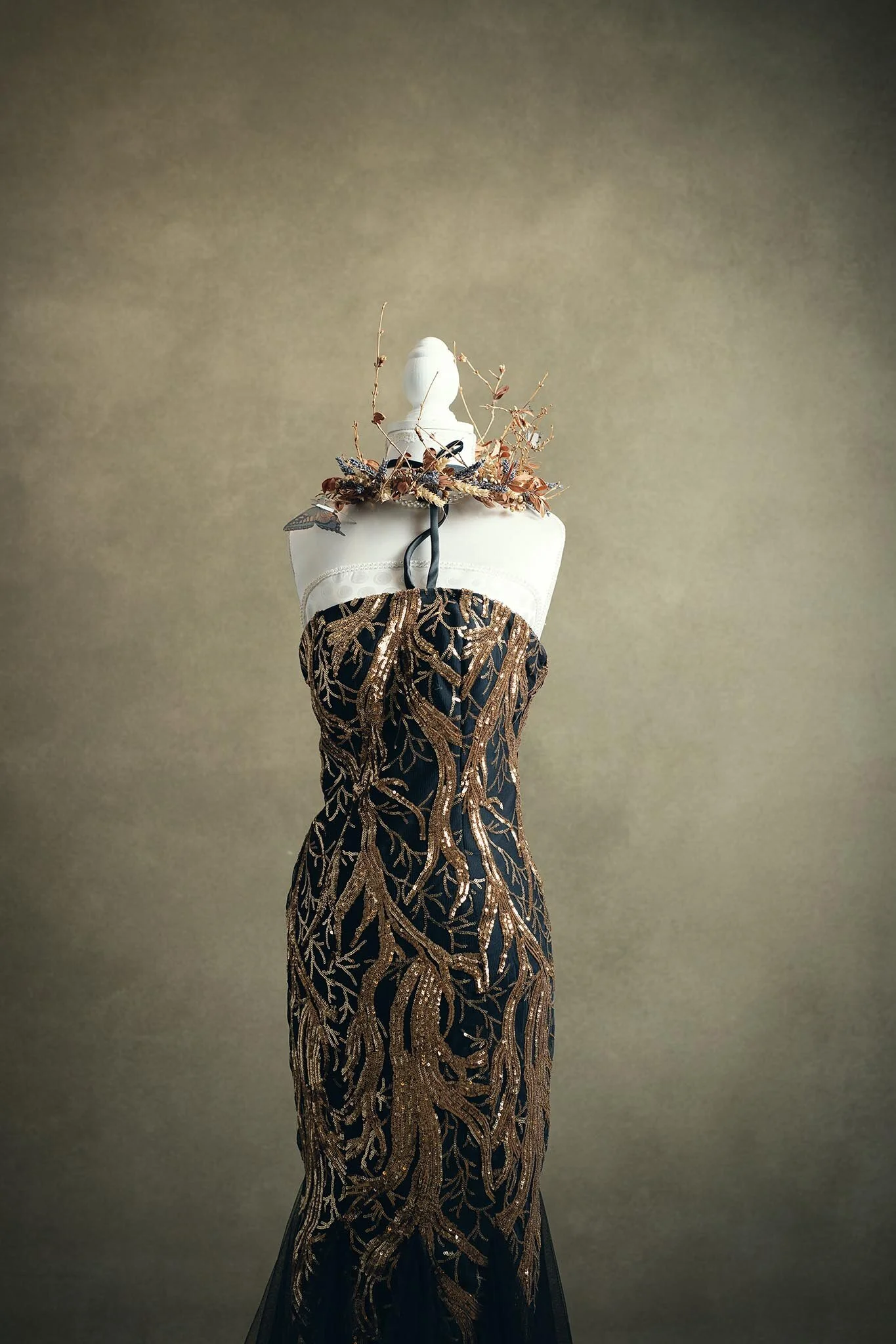 Elegant black dress with intricate gold embroidery displayed on a white mannequin, adorned with a floral necklace made of dried branches and flowers, against a plain beige background.