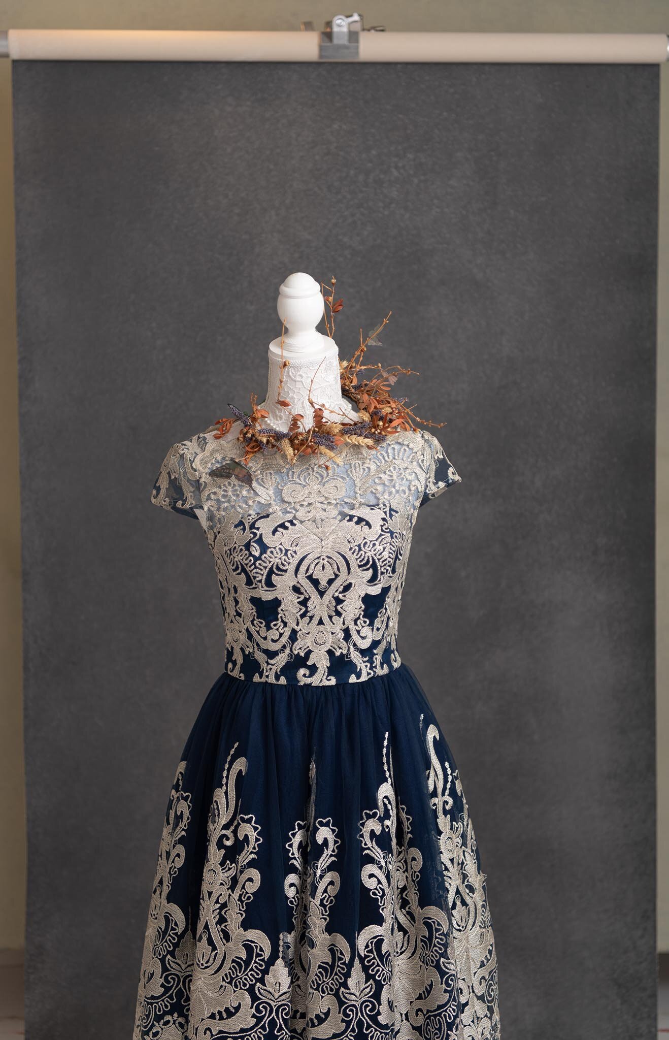 Elegant dress on mannequin, cream lace overlay with navy fabric underneath, intricate floral embroidery, adorned with dried floral jewelry at the neckline, in front of a plain gray background.
