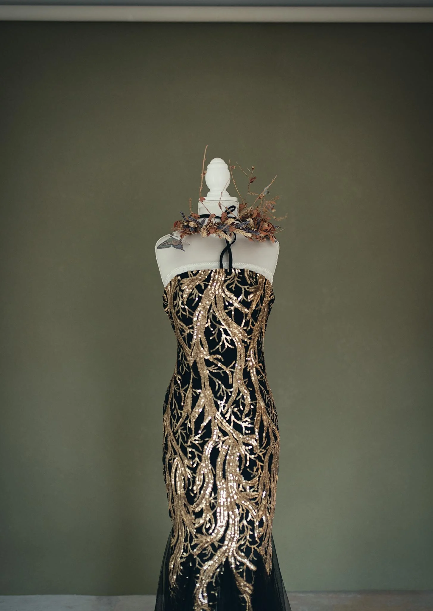 A mannequin dressed in a glamorous black gown with intricate gold embroidery resembling tree branches, placed against a plain wall.