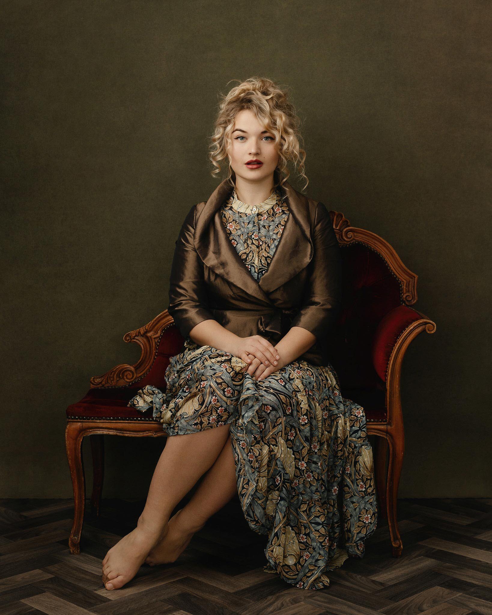 A woman with curly blonde hair sitting on a vintage wooden and red velvet upholstered sofa, wearing a floral dress and a satin bronze-colored blazer, with a dark plain background.