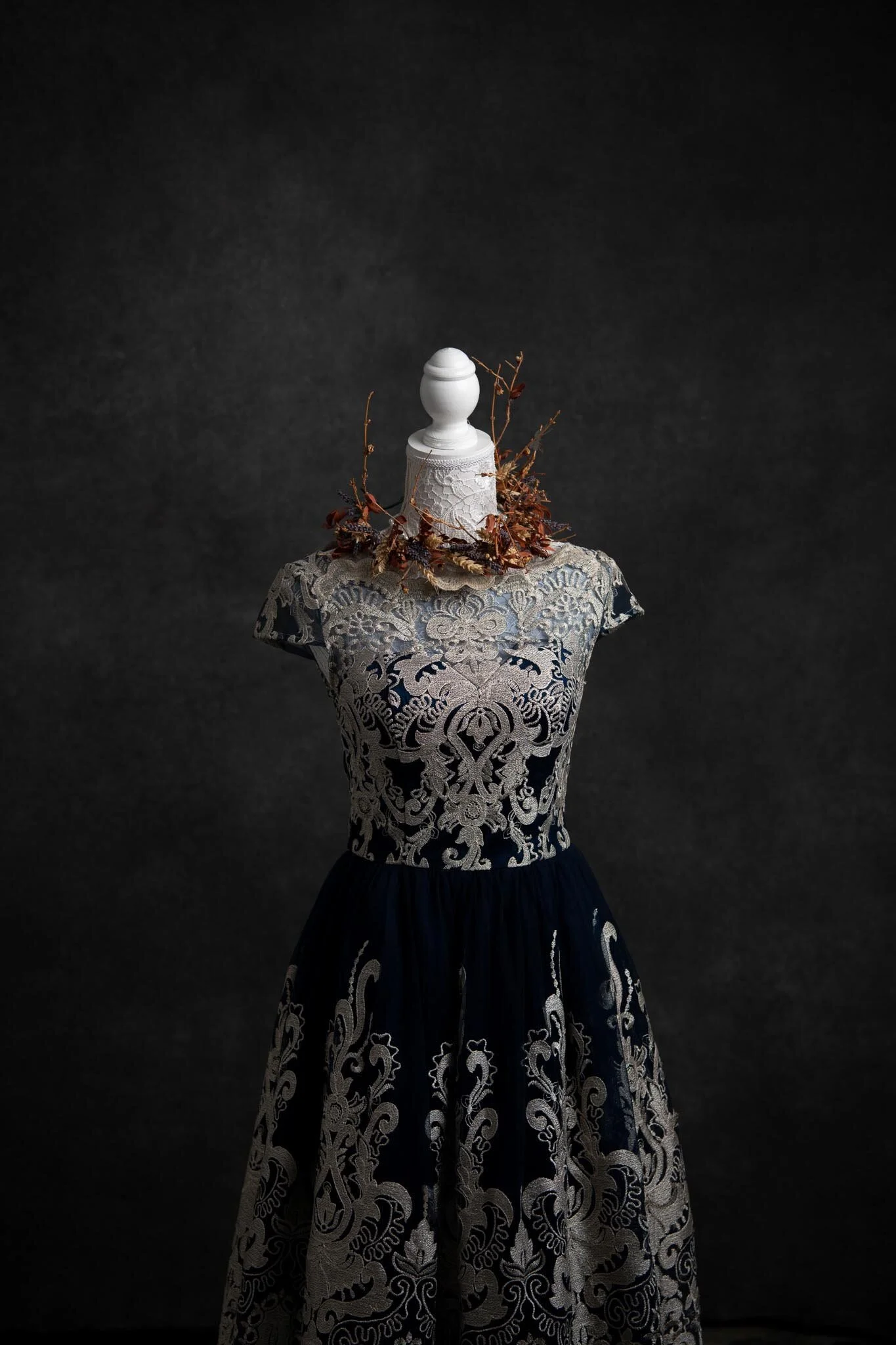 A mannequin dressed in an ornate, black and silver embroidered gown against a dark background, with decorative branches and a white finial on its neck.