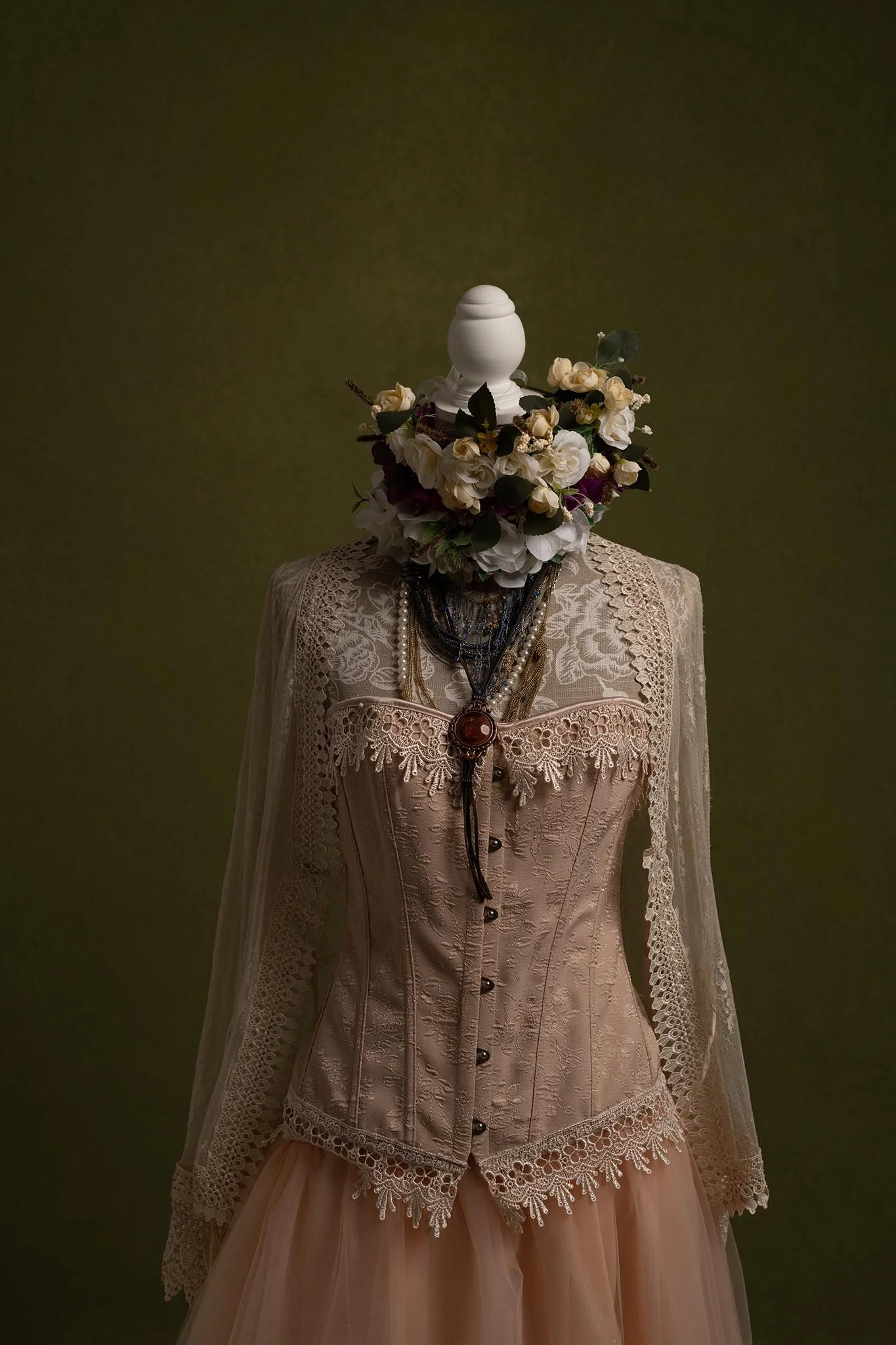 A dress form wearing a vintage lace corset, layered with jewelry, topped with a flower crown made of white and purple flowers, against a plain green background.