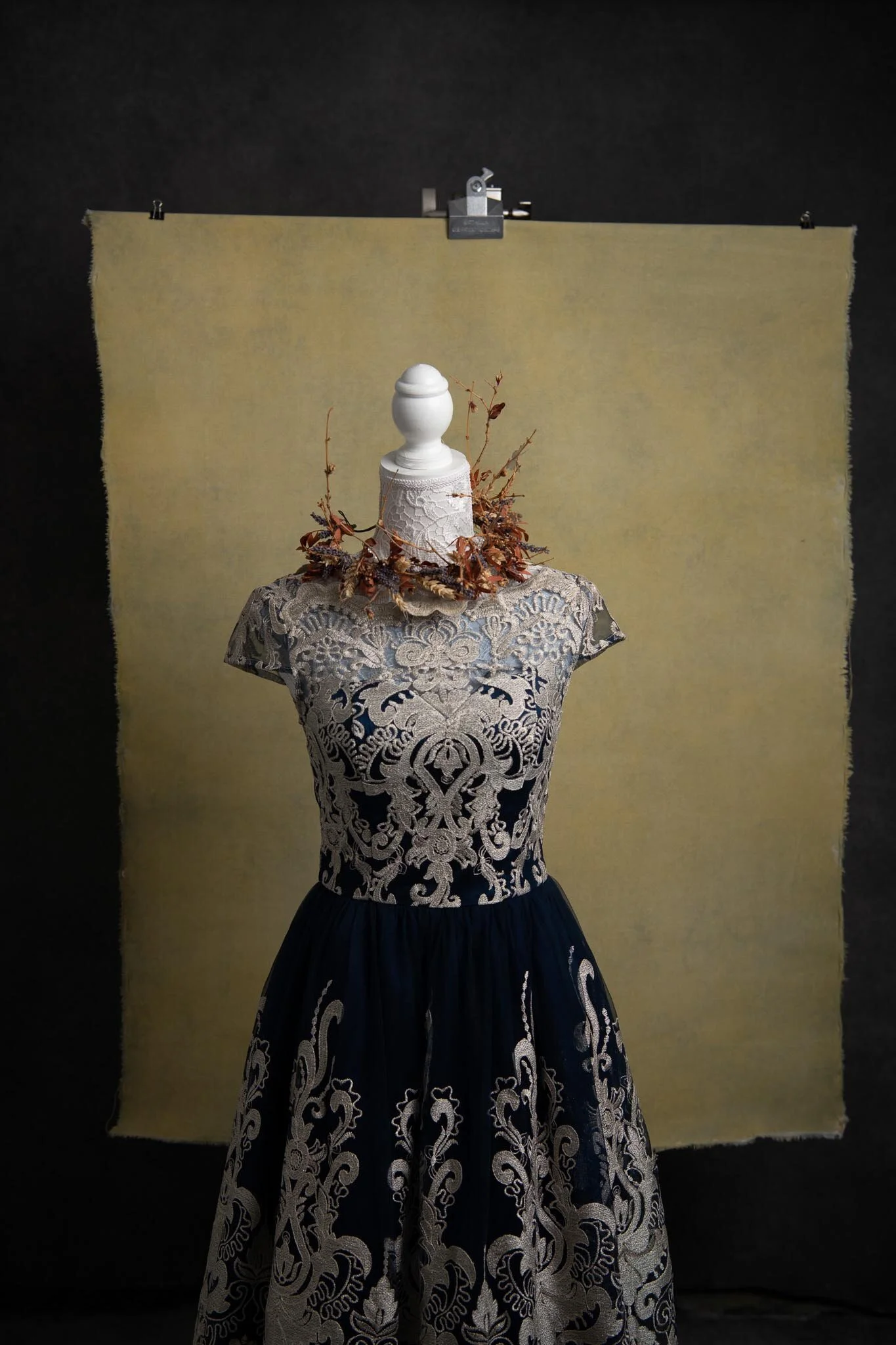 A mannequin wearing a floral and ornamental lace dress with a full skirt, decorated with a wreath of dried branches and leaves around its neck, set against a black and beige backdrop.