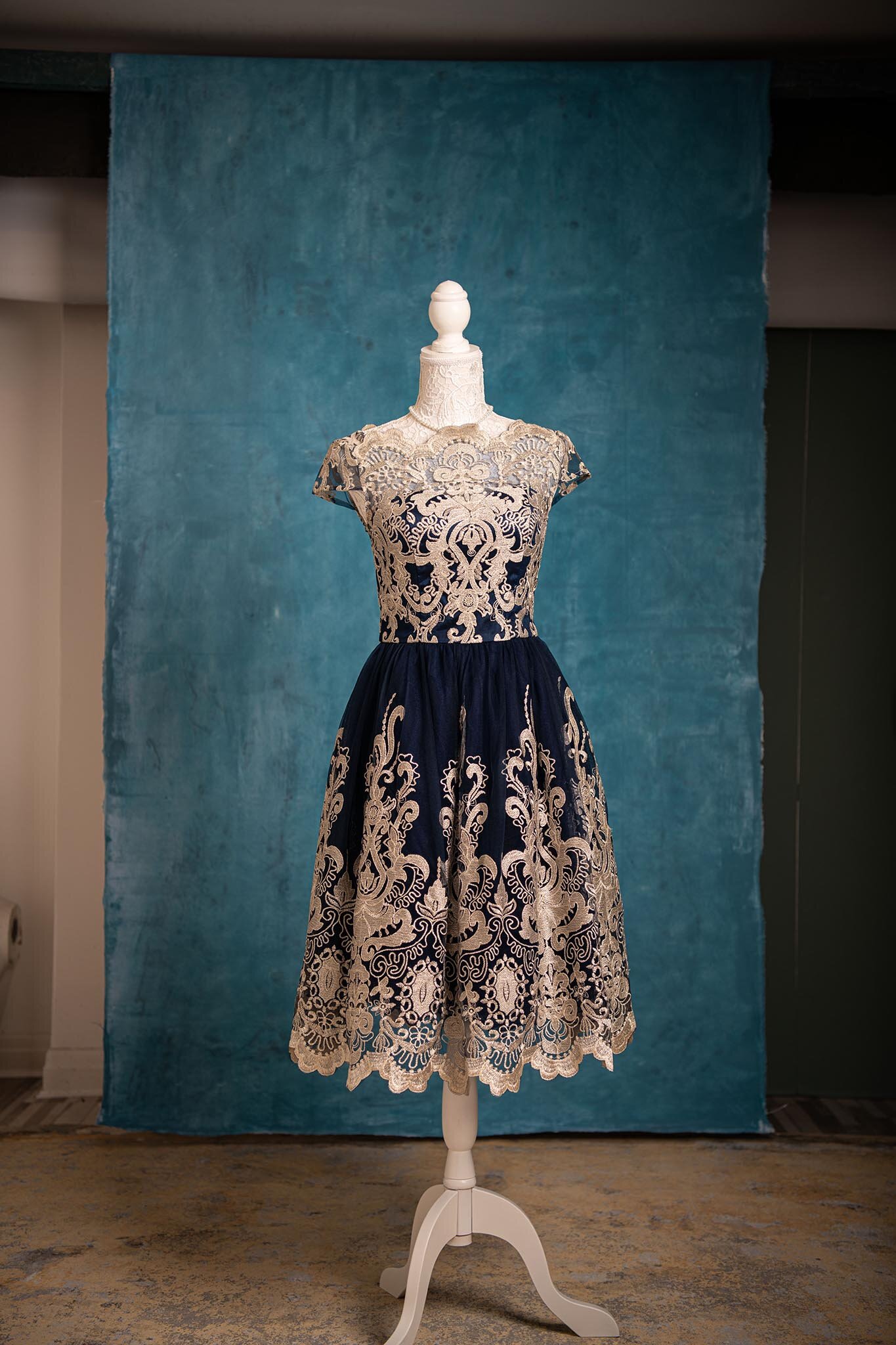 A vintage dress displayed on a mannequin with lace detailing in navy blue and white, set against a textured blue backdrop.