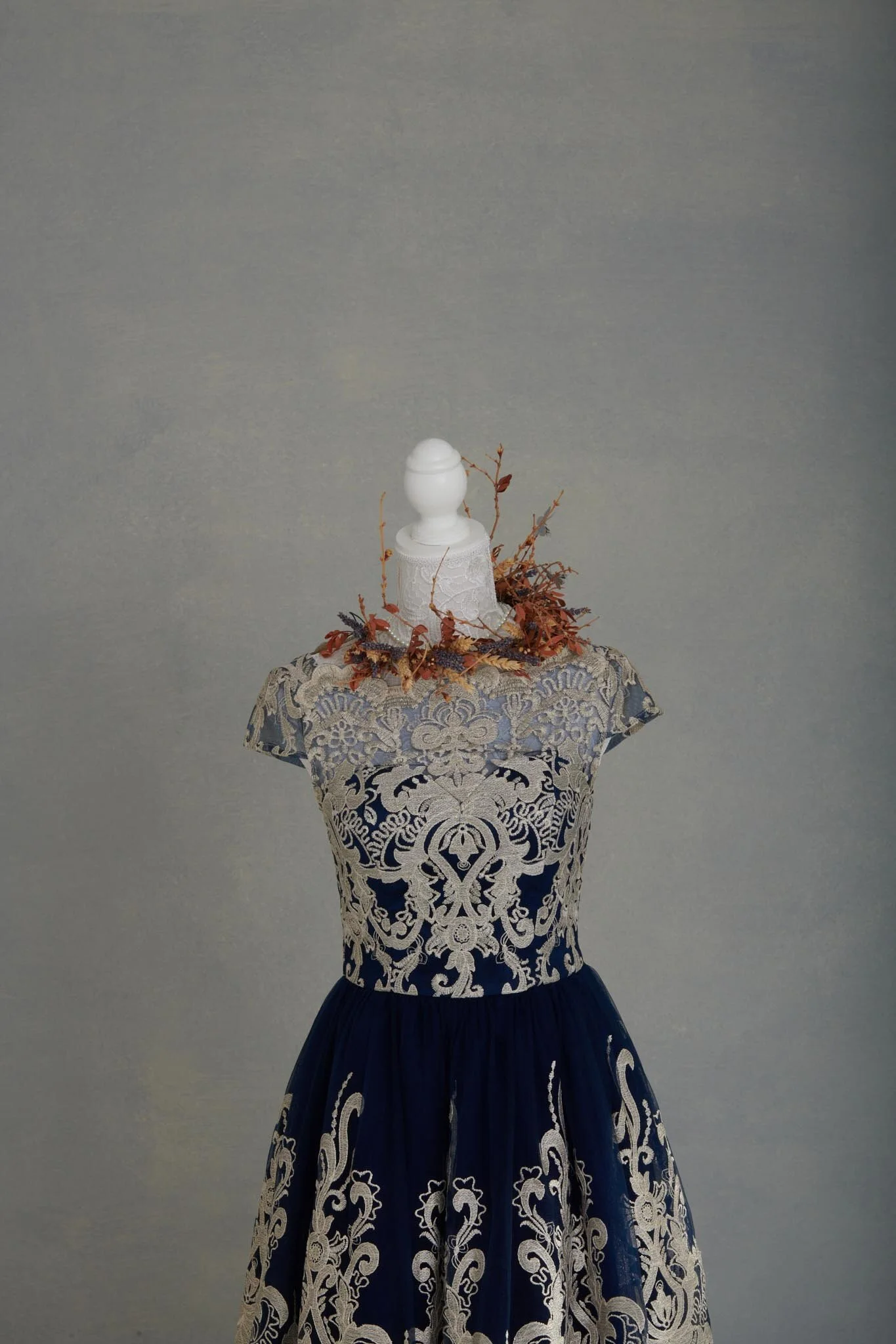 A mannequin wearing a navy blue dress with intricate white lace-like designs, topped with a lace fabric and a decorative piece with autumn leaves, against a plain gray background.