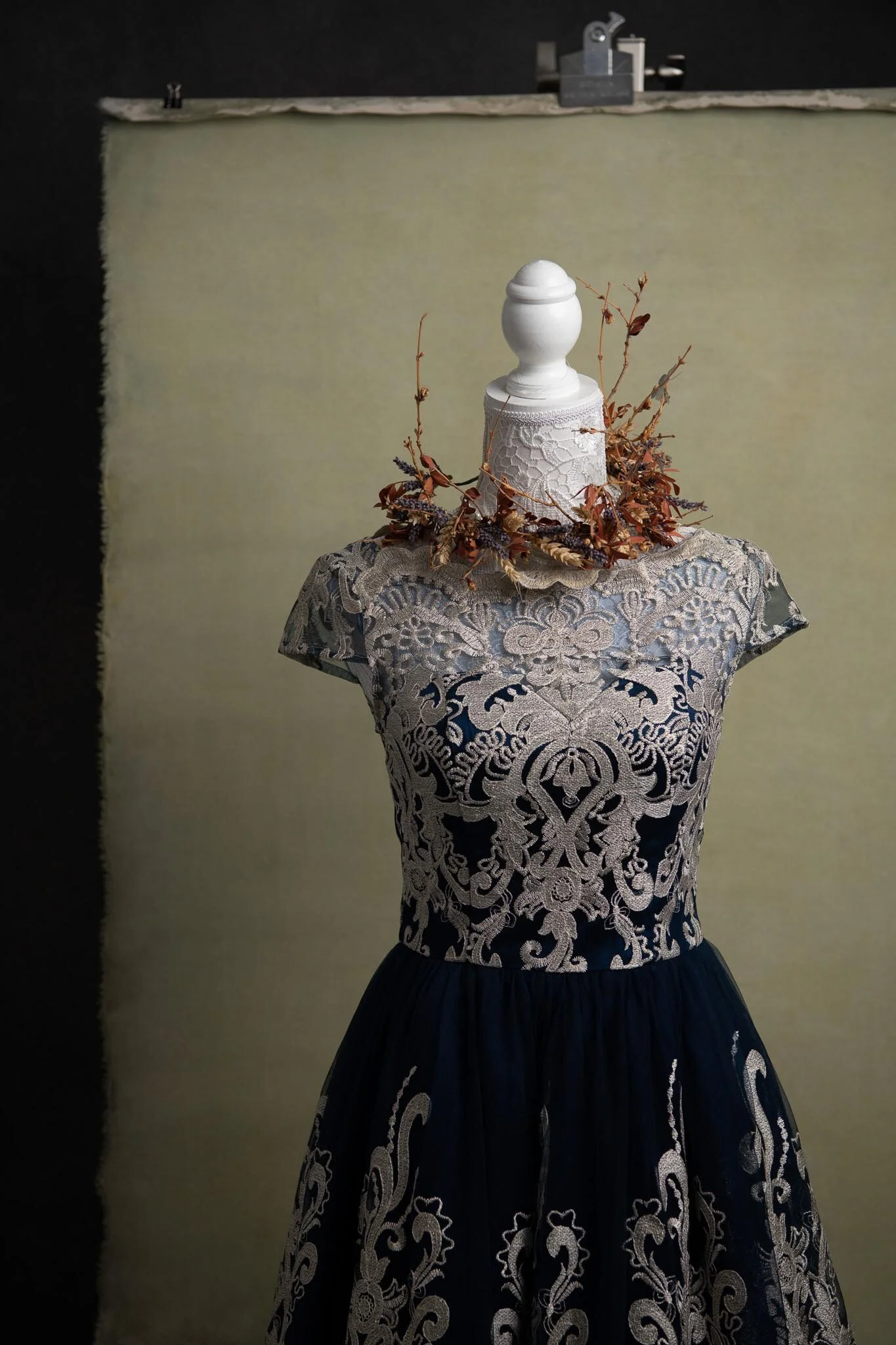 A dress displayed on a mannequin with a floral wreath around its neck, set against a pale background with a black and a metallic item on a shelf at the top.