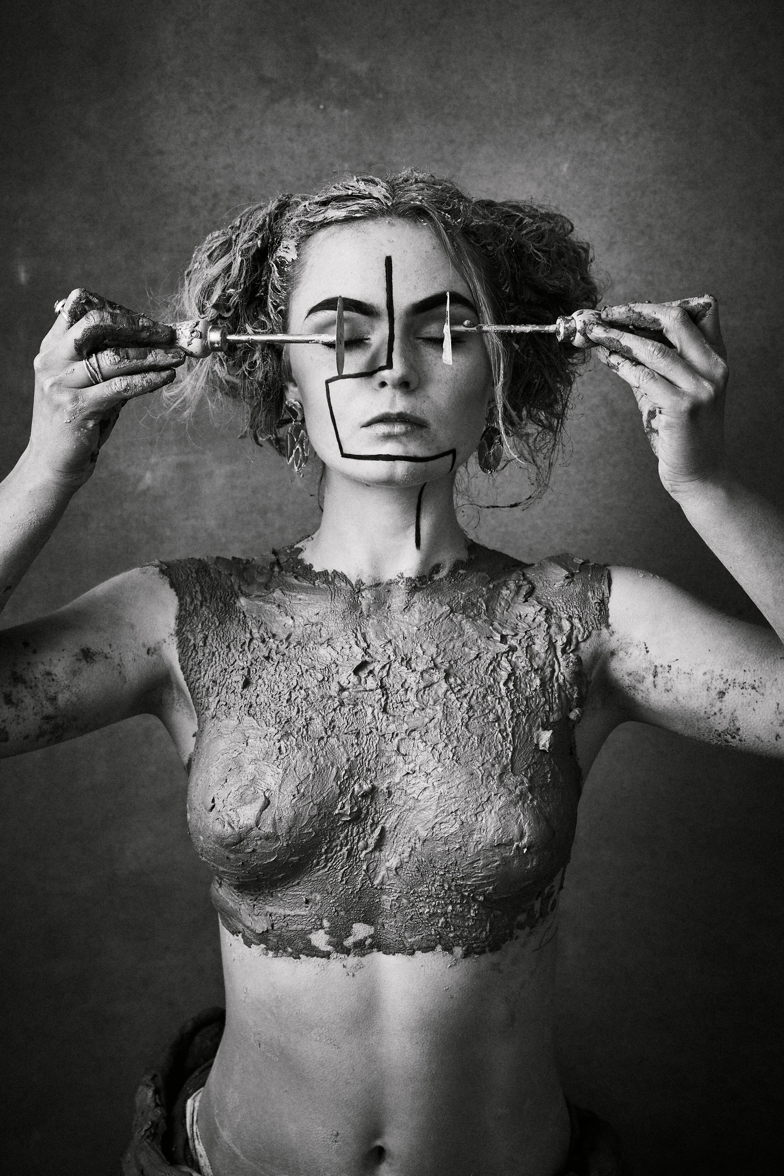 Black and white artistic portrait of Mira - woman with paint brushes held across her face, against a textured grey hand painted canvas backdrop by Hand Painted Backdrops UK.