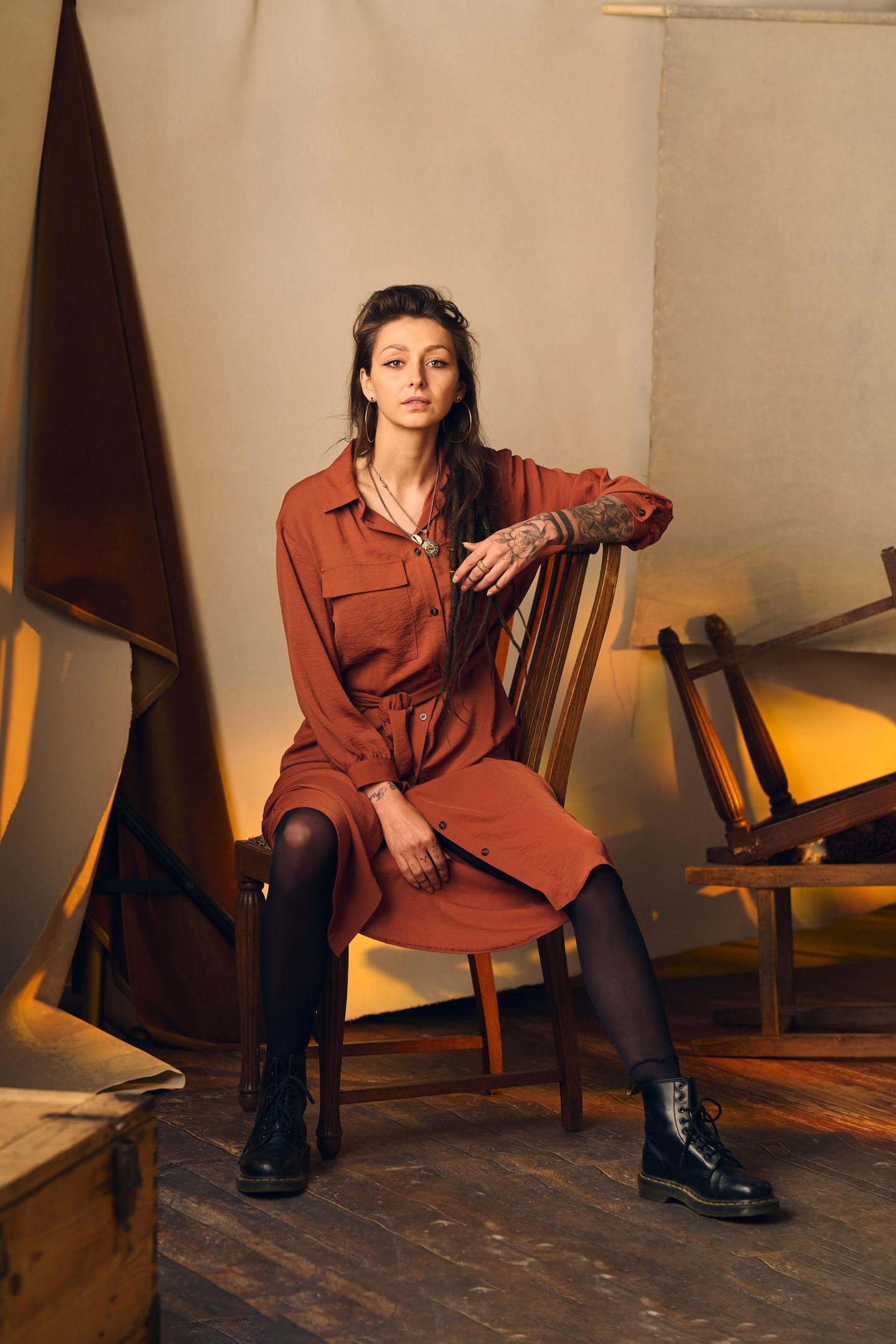 That soft strategic lighting is washing over this scene creating natural warmth that makes her rust-colored dress absolutely sing against the muted beige hand-painted backdrop while highlighting every detail from the fabric texture to those intricate