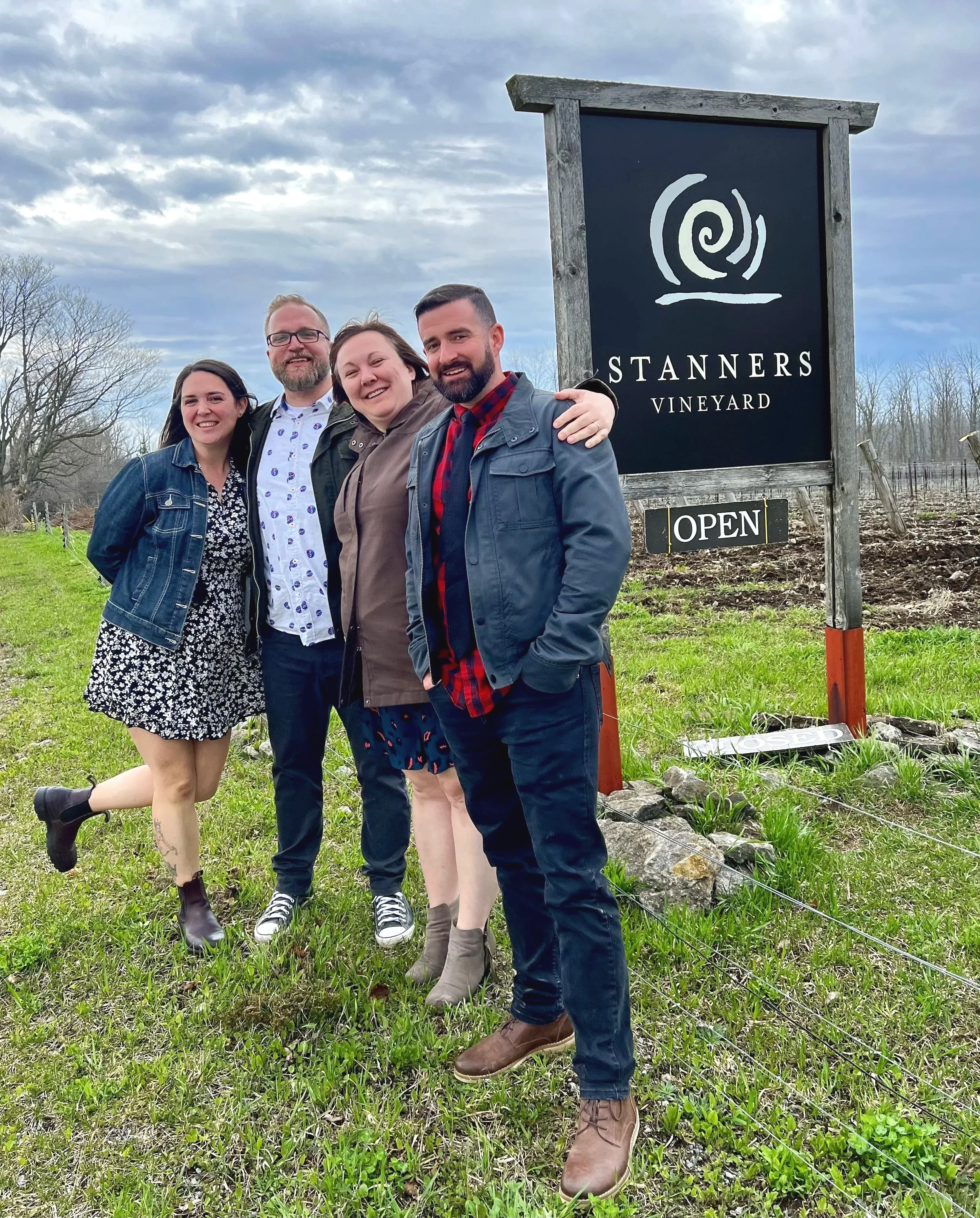 Four people standing together outdoors in front of a sign for Stanners Vineyard, smiling and posing for a photo.