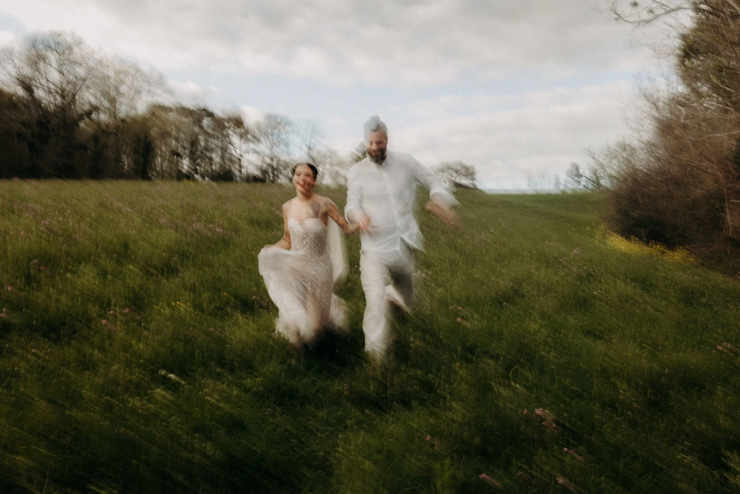 Un couple en mariage, la femme en robe blanche et l'homme en costume blanc, court joyeusement dans une prairie verte avec des arbres en arrière-plan, sous un ciel partiellement nuageux.