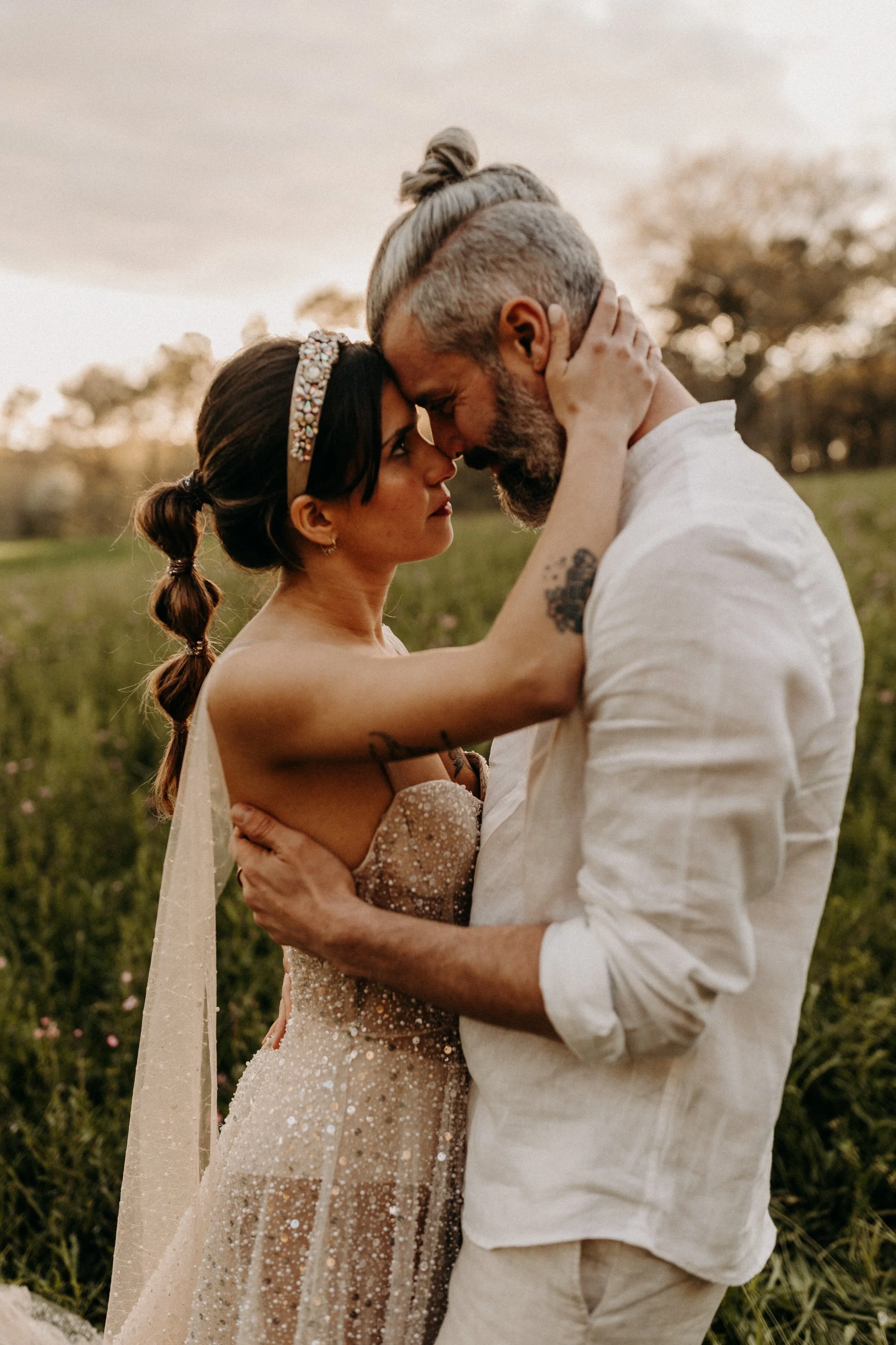 Un couple s'embrasse dans un champ lors d'un coucher de soleil, l'homme porte une chemise blanche, la femme porte une robe de mariée élégante avec un bandeau décoratif dans les cheveux.