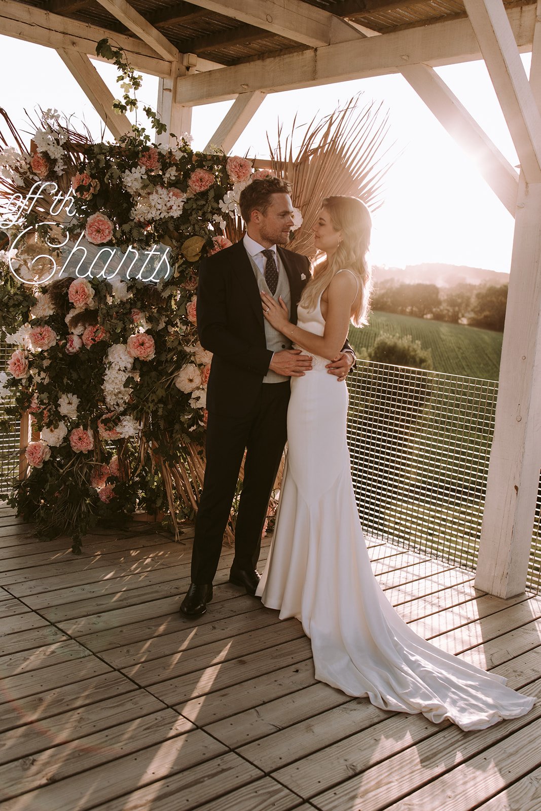 Un couple habillé de manière élégante, l'homme en costume et la femme en robe blanche, se tient sous une structure en bois devant un arrangement floral arborant une inscription lumineuse. La scène est illuminée par la lumière du coucher du soleil.