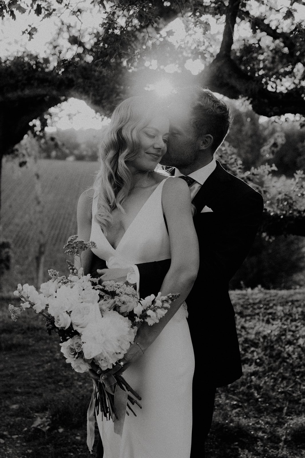 Un couple romantique en tenue de mariage, la femme tenant un bouquet de fleurs, dans un cadre extérieur avec des arbres en arrière-plan.