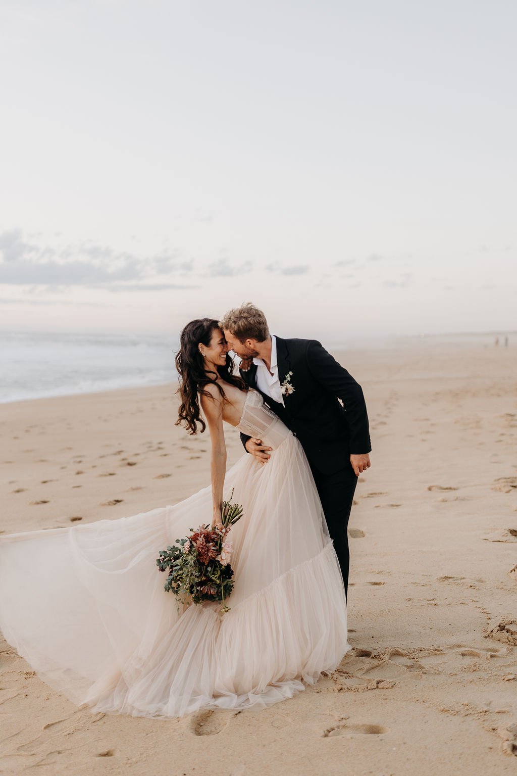 Un couple en tenue de mariage s'embrasse sur une plage, la mariée tenant un bouquet de fleurs. Ils sont entourés de sable et l'océan est visible en arrière-plan.