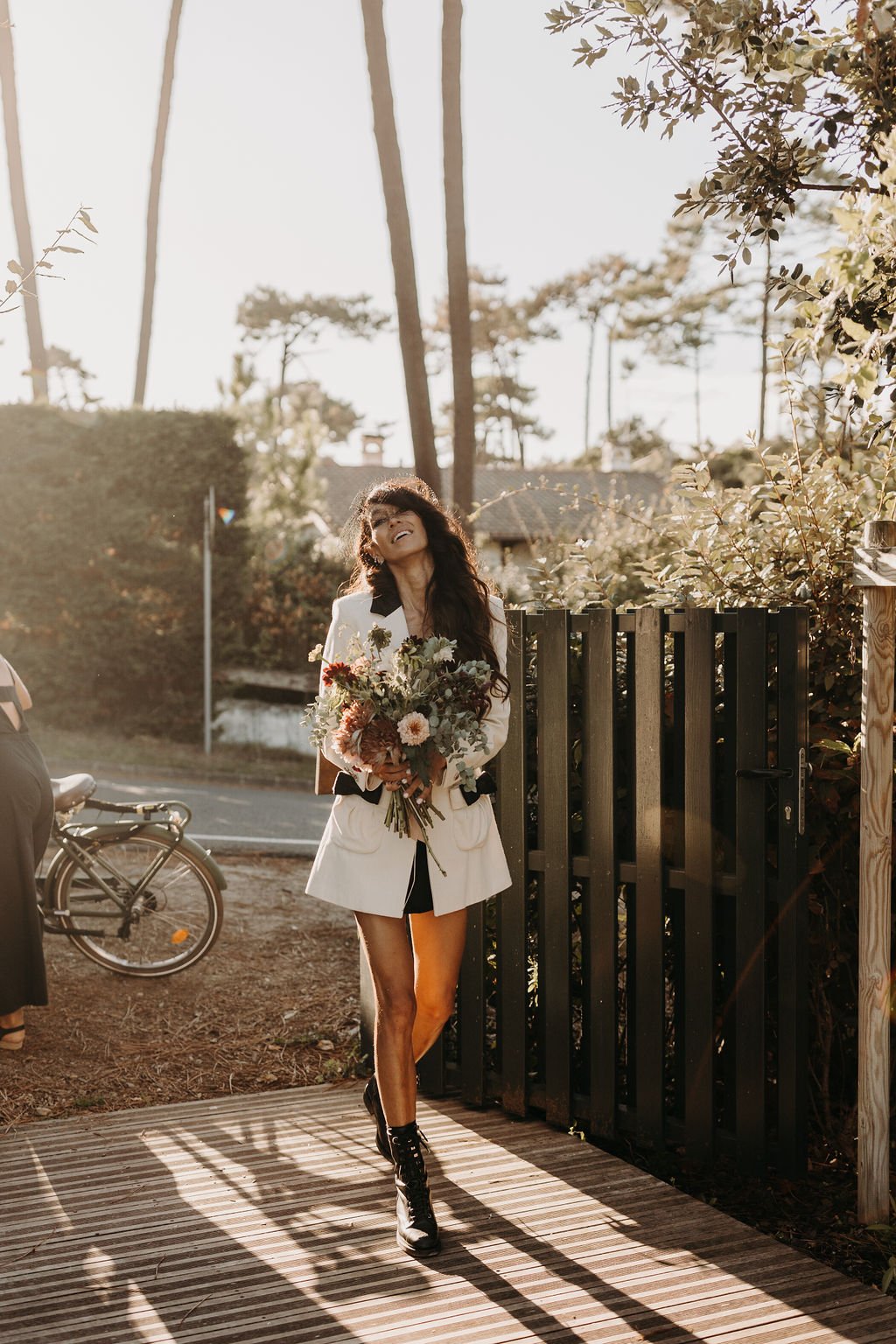 Femme souriante tenant un bouquet de fleurs, marchant sur une terrasse en bois avec une barrière et des arbres en arrière-plan.