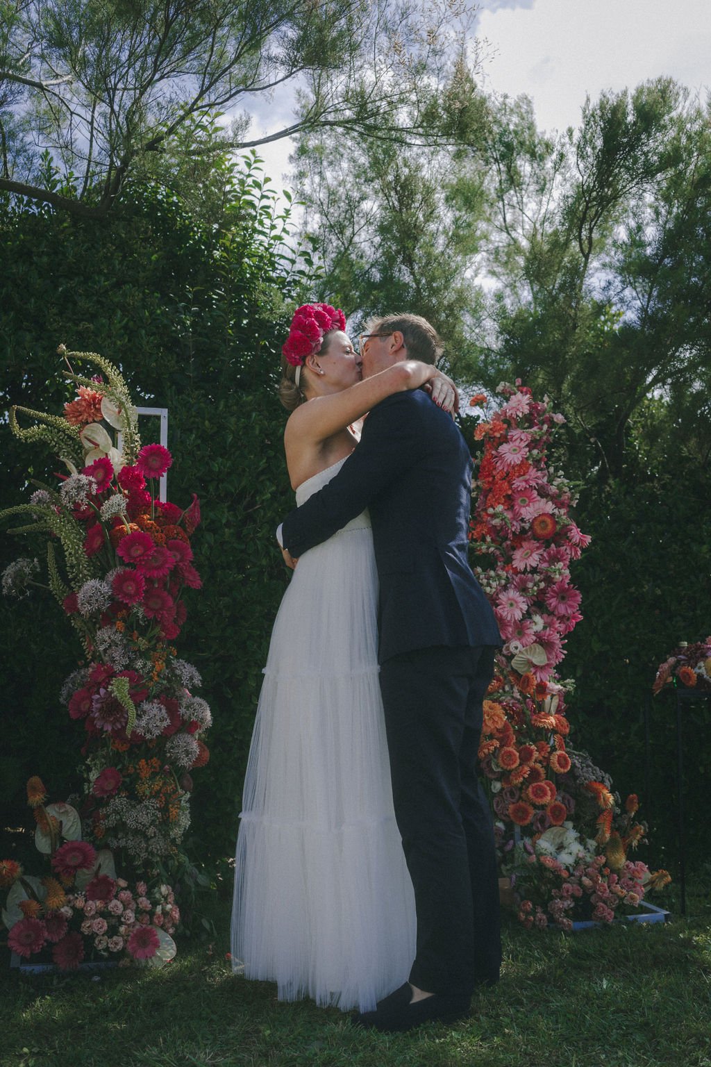 Un couple s'embrasse lors d'une cérémonie de mariage. La mariée porte une robe blanche et un diadème de fleurs rouges, tandis que le marié est en costume noir. Ils sont entourés de compositions florales colorées.