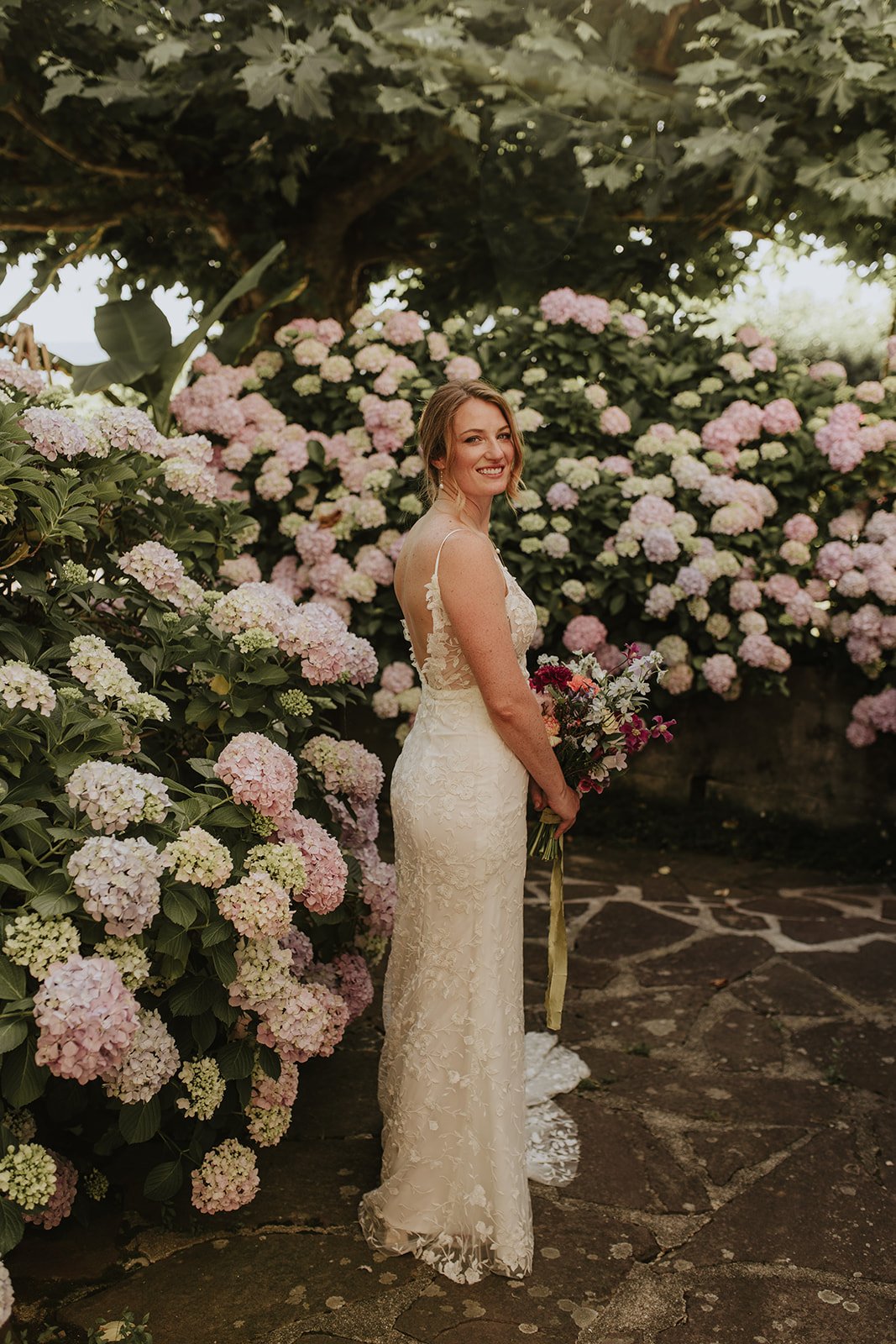 Une femme en robe de mariée debout près d'arbustes fleuris avec des hortensias roses et blancs, tenant un bouquet de fleurs colorées.