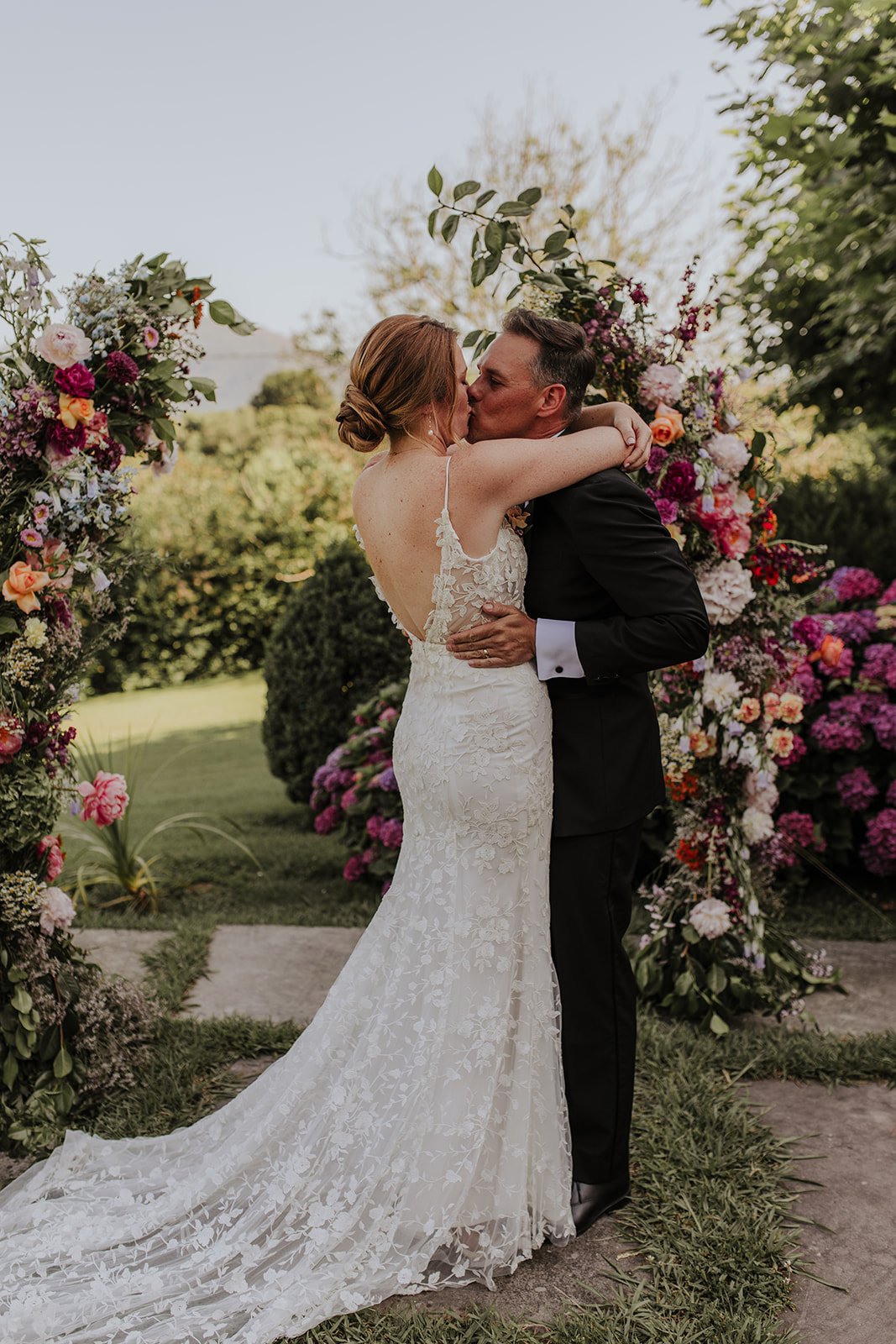 Un couple de mariés s'embrassant lors d'une cérémonie en plein air, entouré de compositions florales.