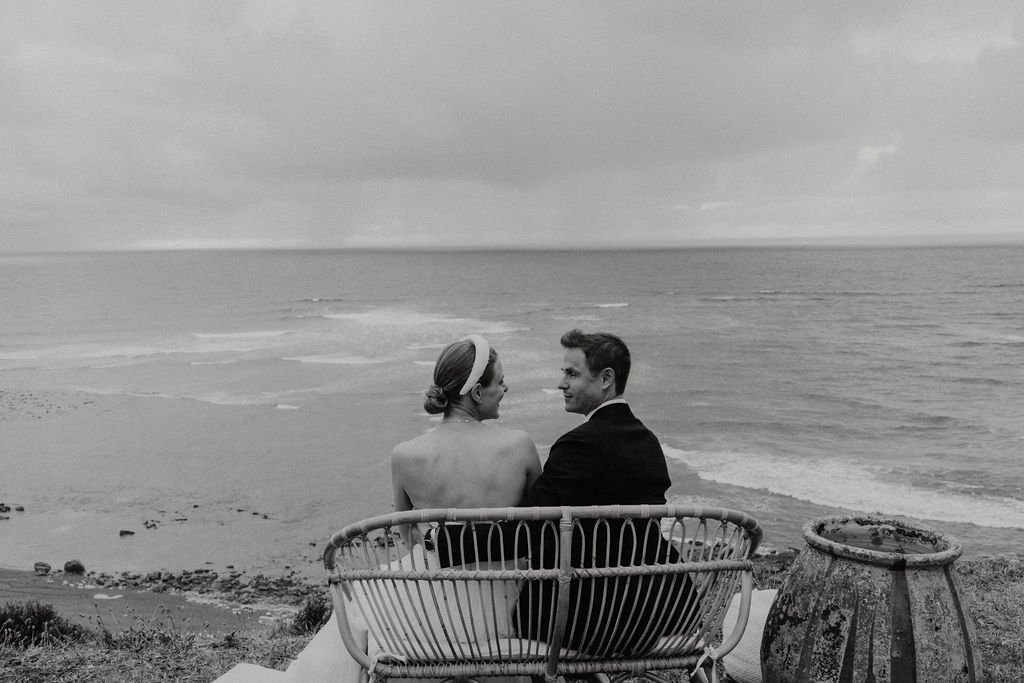 Un couple assis sur un banc en osier fait face à la mer, regardant l'horizon. L'image est en noir et blanc, créant une ambiance romantique et apaisante.