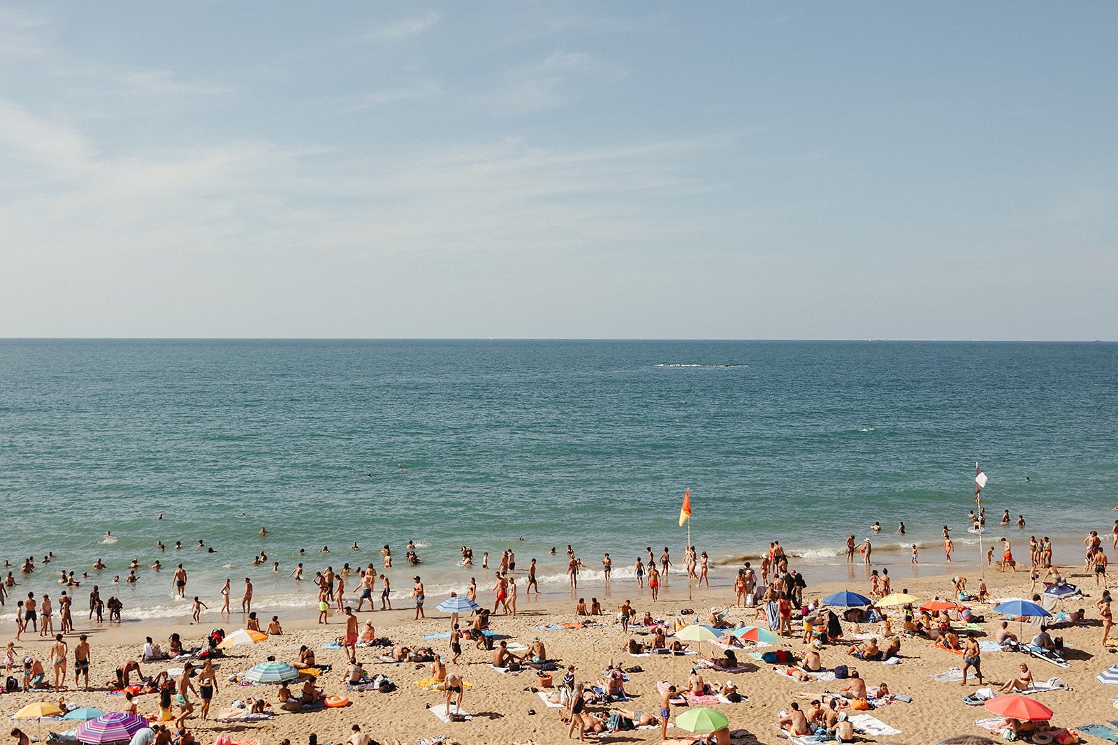 Plage bondée avec des gens se relaxant sur le sable et nageant dans la mer. Des parasols multicolores sont ouverts. Ciel bleu au-dessus.