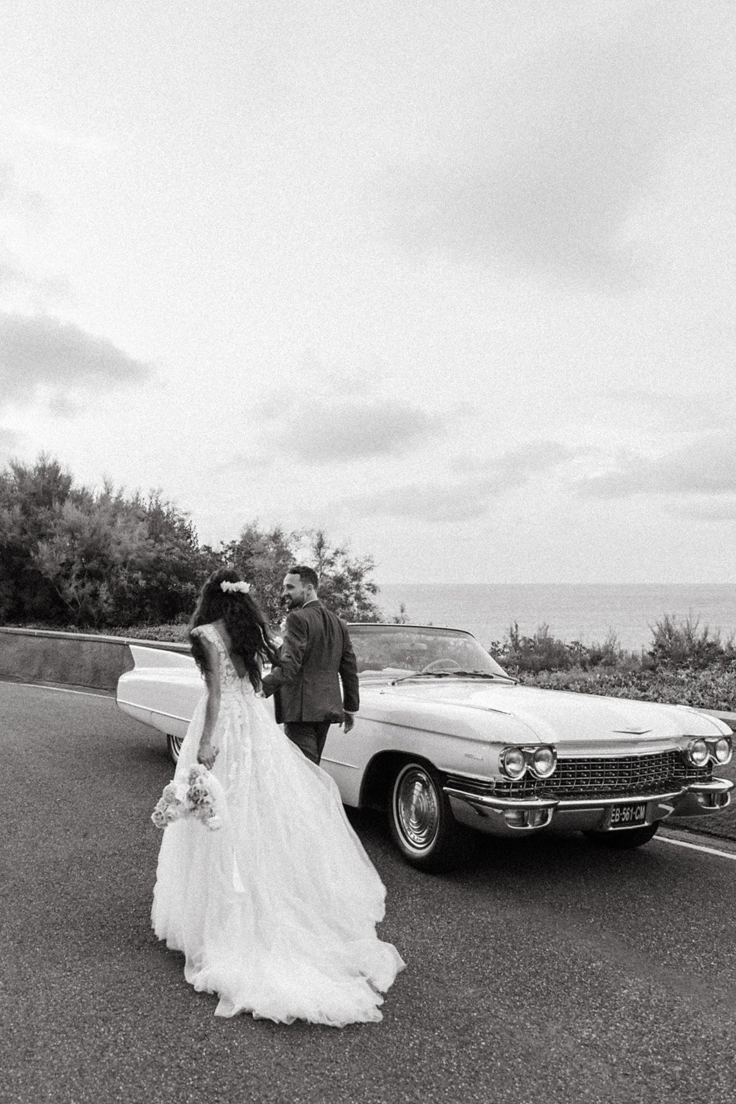 Un couple en tenue de mariage marche près d'une voiture classique sur une route, avec la mer en arrière-plan.