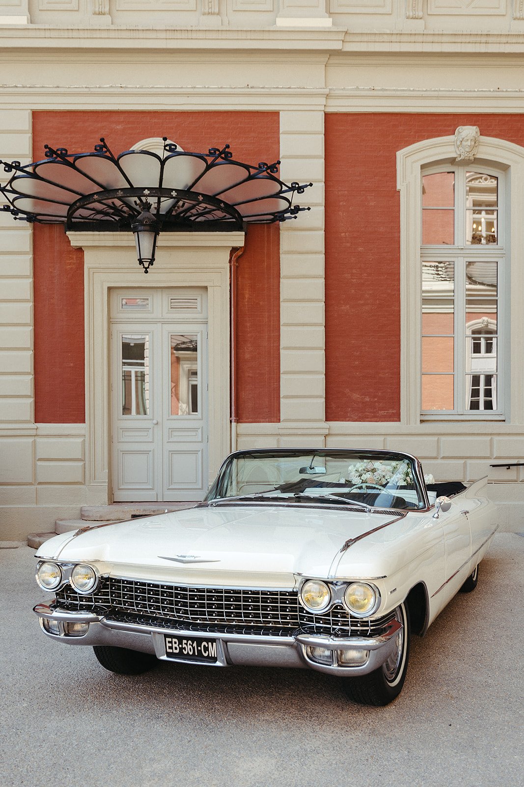 Voiture décapotable vintage blanche devant une façade de bâtiment rouge et beige.