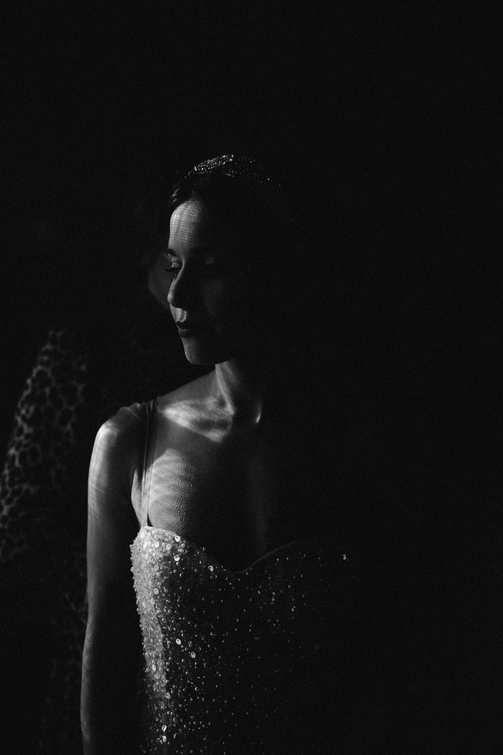 Une femme en robe ornée de sequins, photographiée en noir et blanc dans une faible lumière, créant un contraste dramatique.