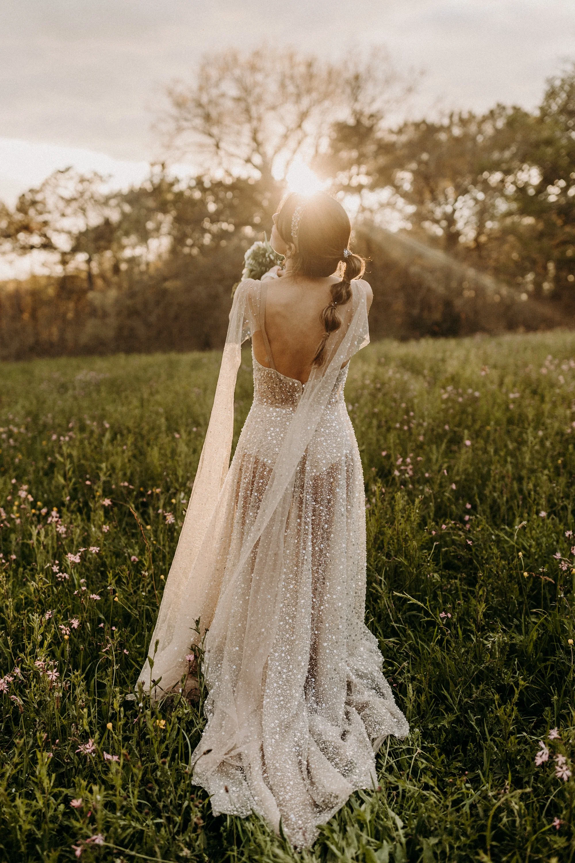 Une femme en robe de mariage élégante, vue de dos, dans un champ de fleurs pendant le coucher de soleil.
