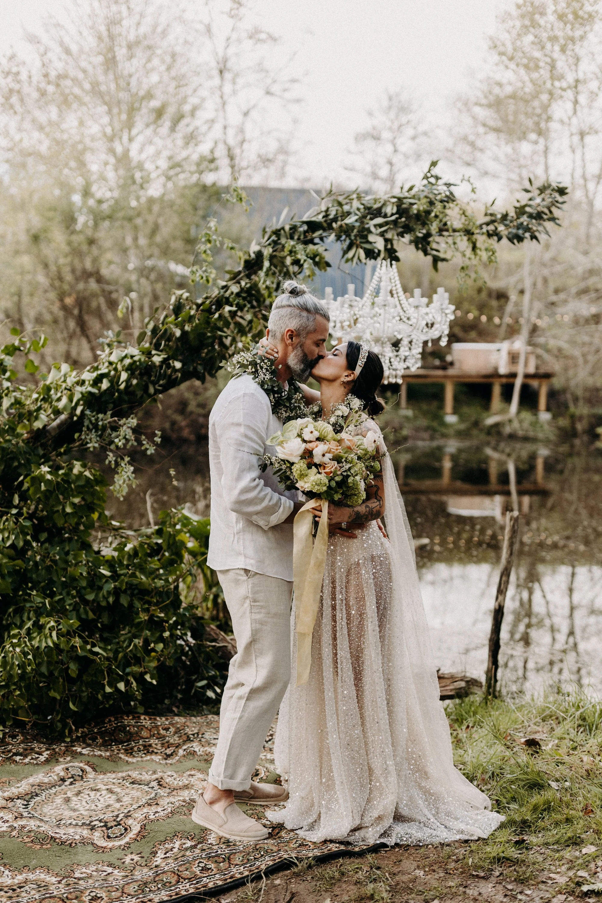 Un couple lors de leur mariage en plein air, s'embrassant avec un bouquet de fleurs, sous une arche décorée de feuillage et d'une grande chandelier, près d'un étang.