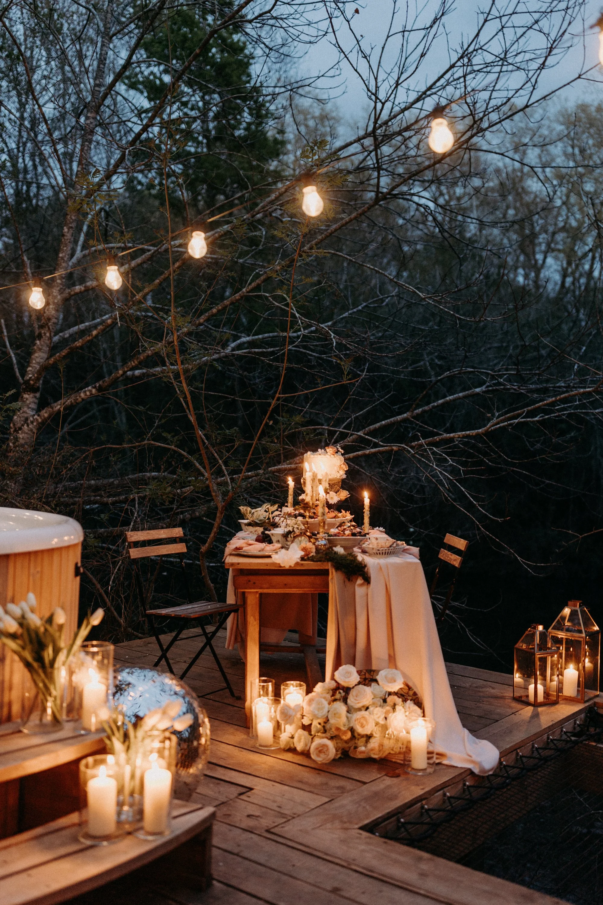 Table décorée pour un événement en plein air, avec bougies, fleurs blanches, guirlande lumineuse, et lanternes en verre, dans un environnement boisé au crépuscule.