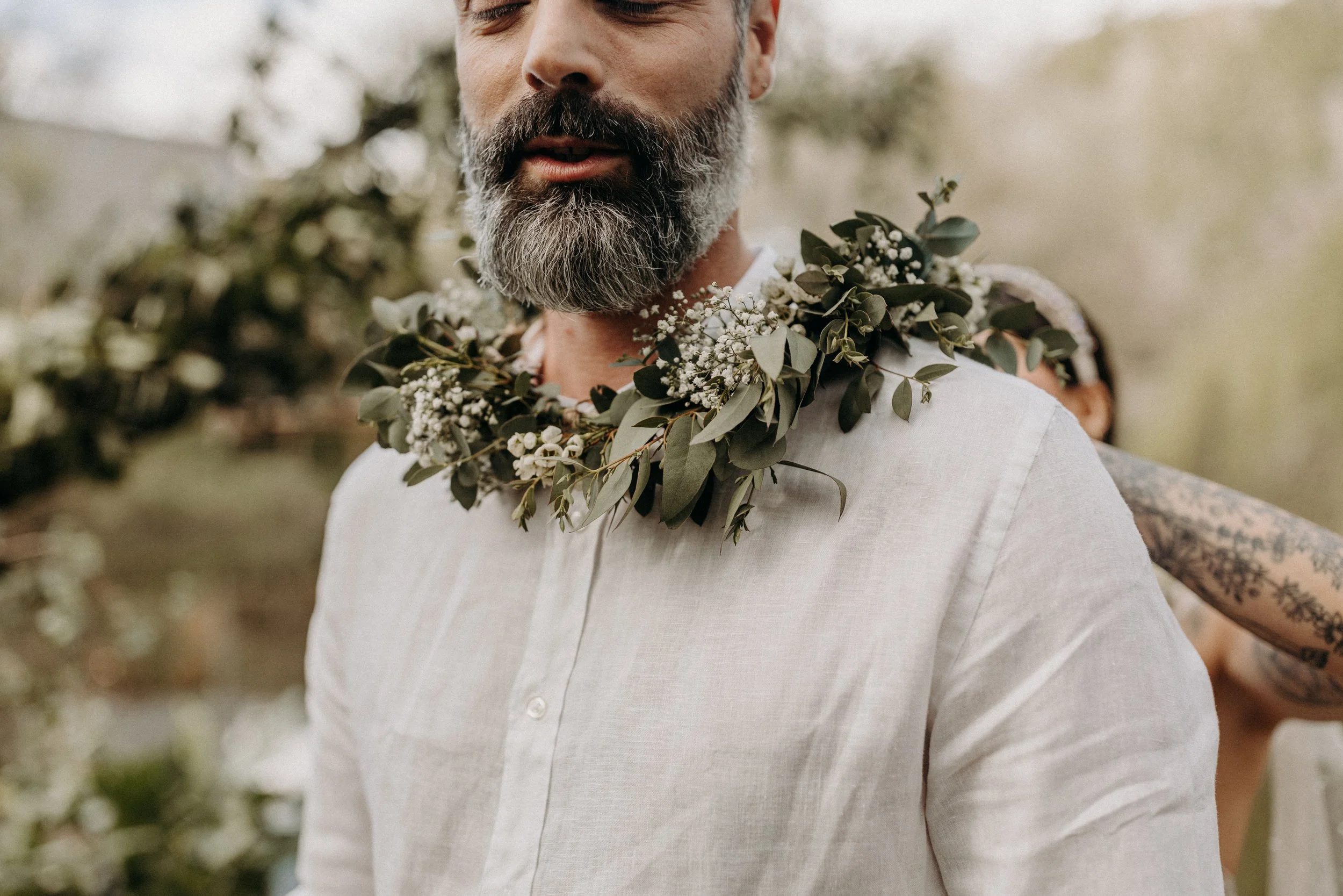 Homme avec barbe portant une chemise blanche et un collier de branches et fleurs blanches, en extérieur.