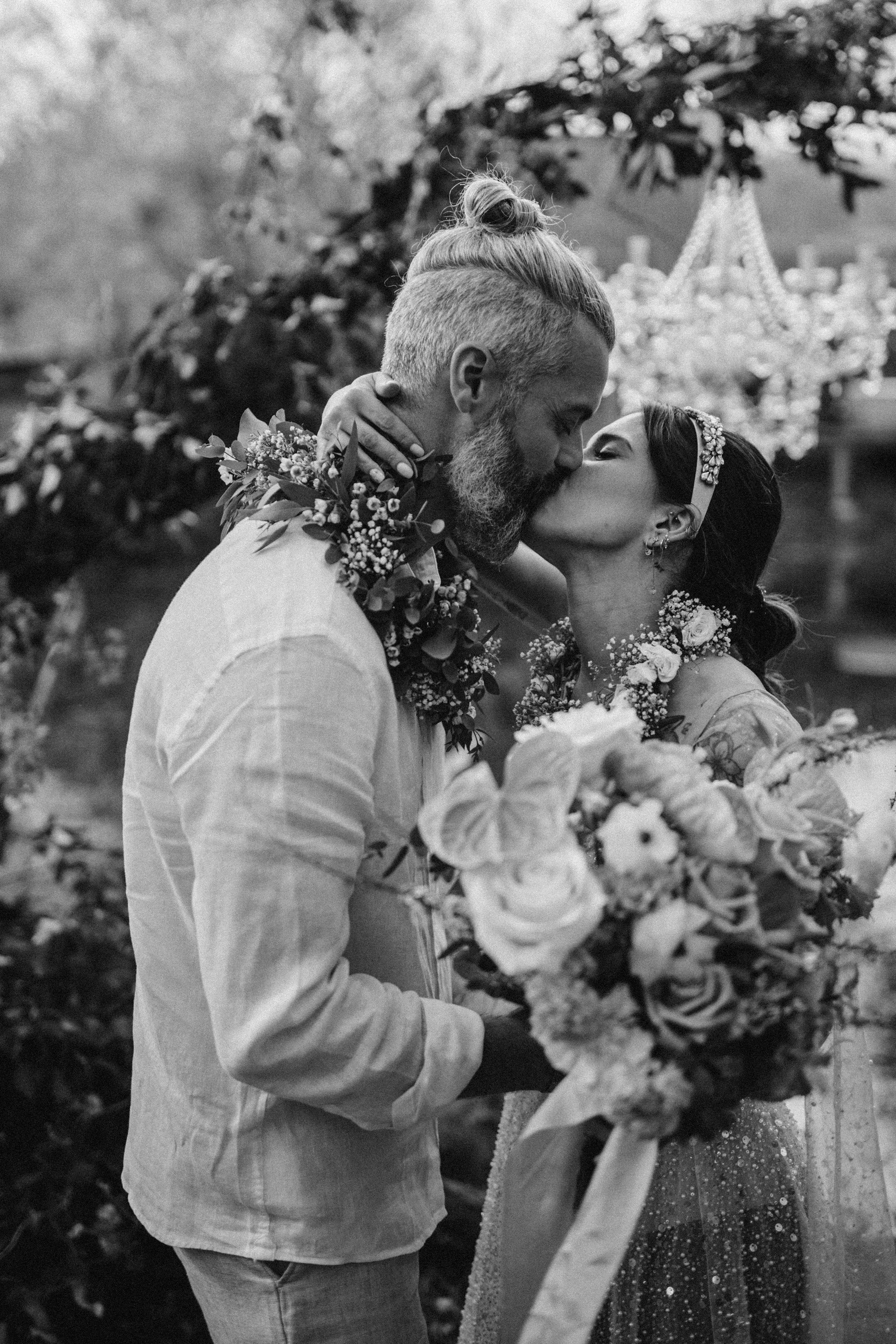 Un couple de mariés s'embrassent lors de leur mariage, entourés de fleurs, dans une photographie en noir et blanc.
