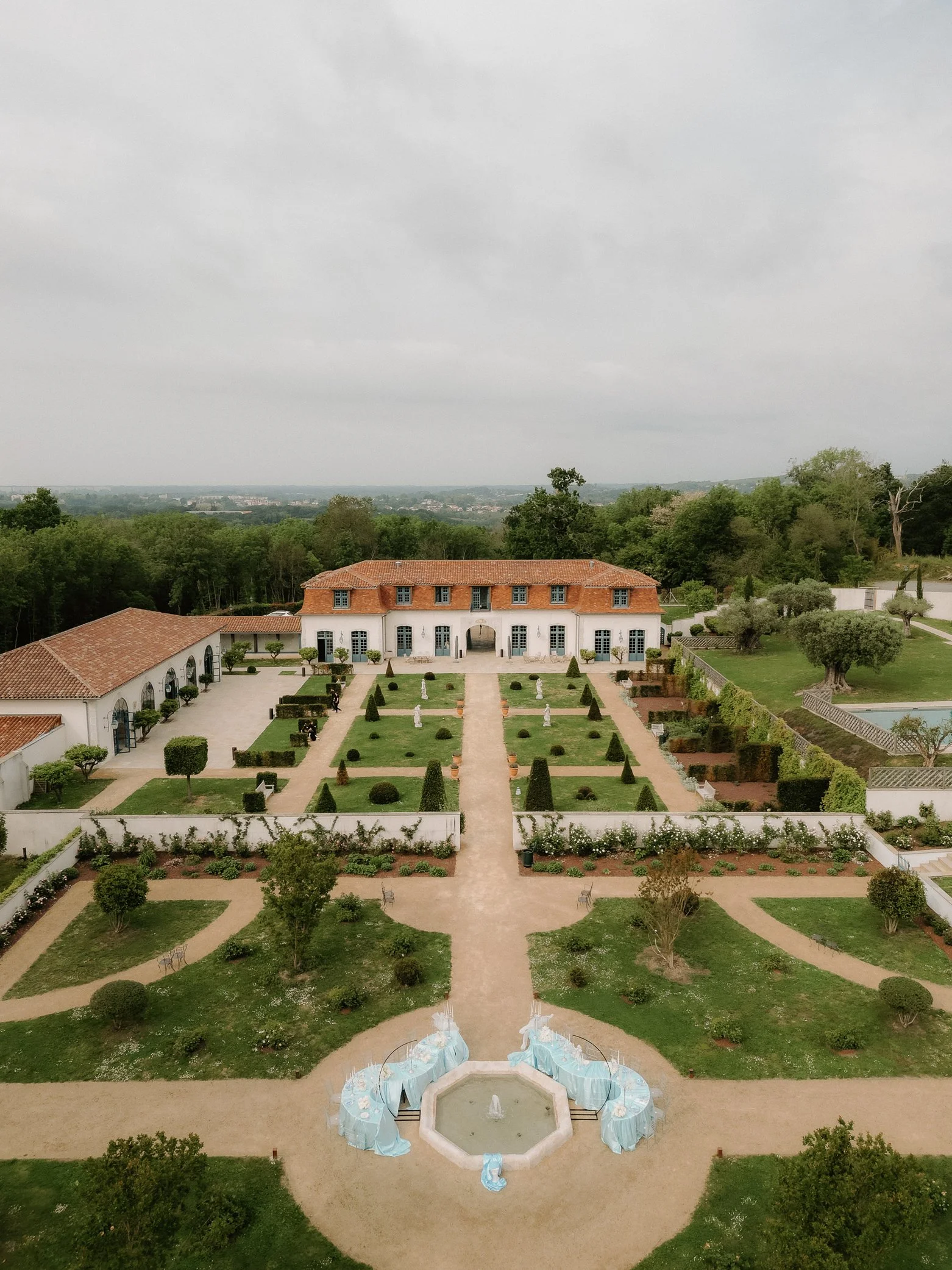 Un manoir avec une façade en pierre blanche et un toit en tuiles rouges, entouré de jardins bien entretenus avec des statues et des pots de fleurs. Des personnes sont rassemblées devant, probablement pour un événement.