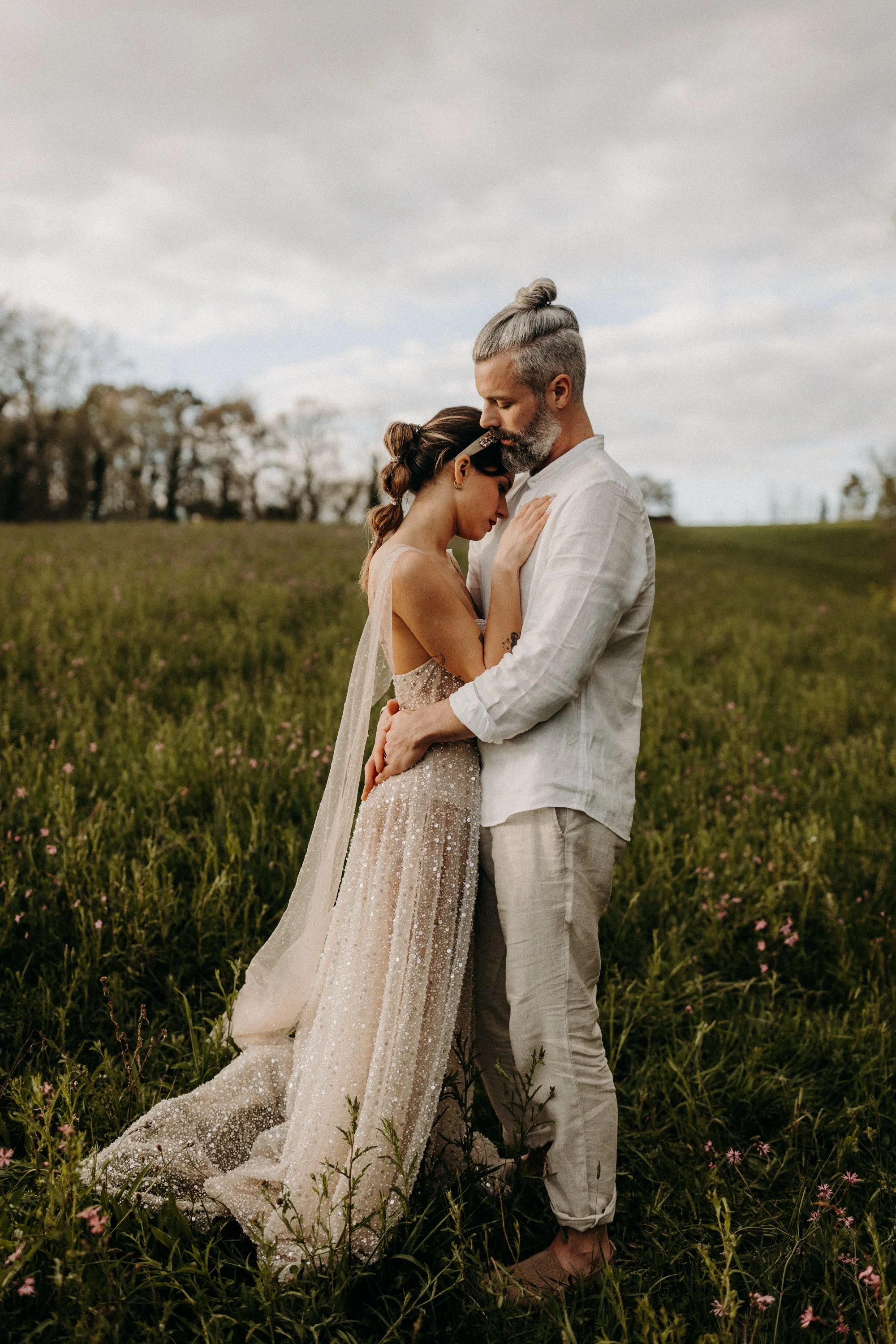 Un couple en habits de mariage dans un champ, se tenant très proches, avec un ciel nuageux en arrière-plan.