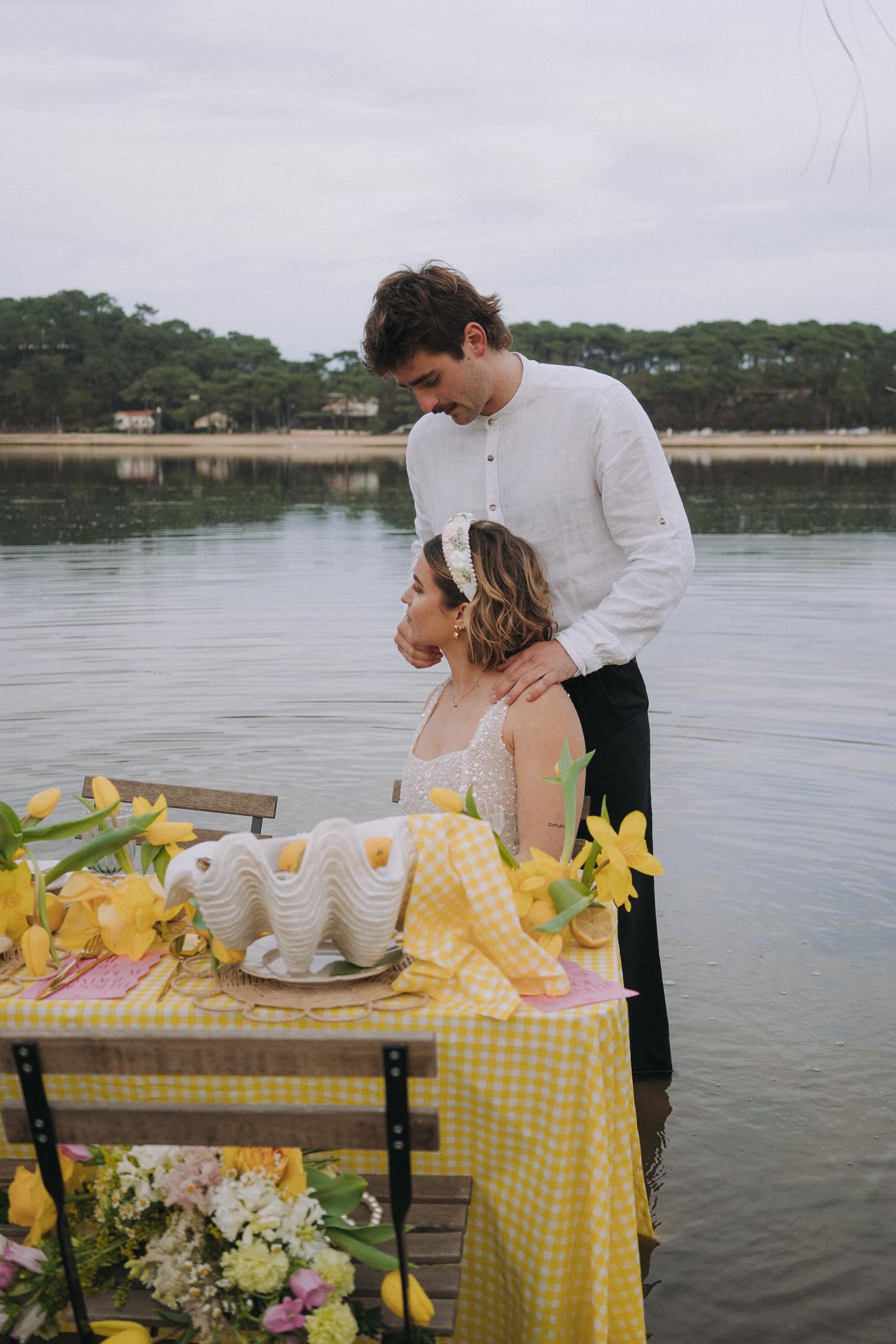 Un couple élégant, avec un homme en chemise blanche, pose près d'une table ornée de fleurs jaunes et d'une nappe à carreaux jaunes, au bord d'un lac.