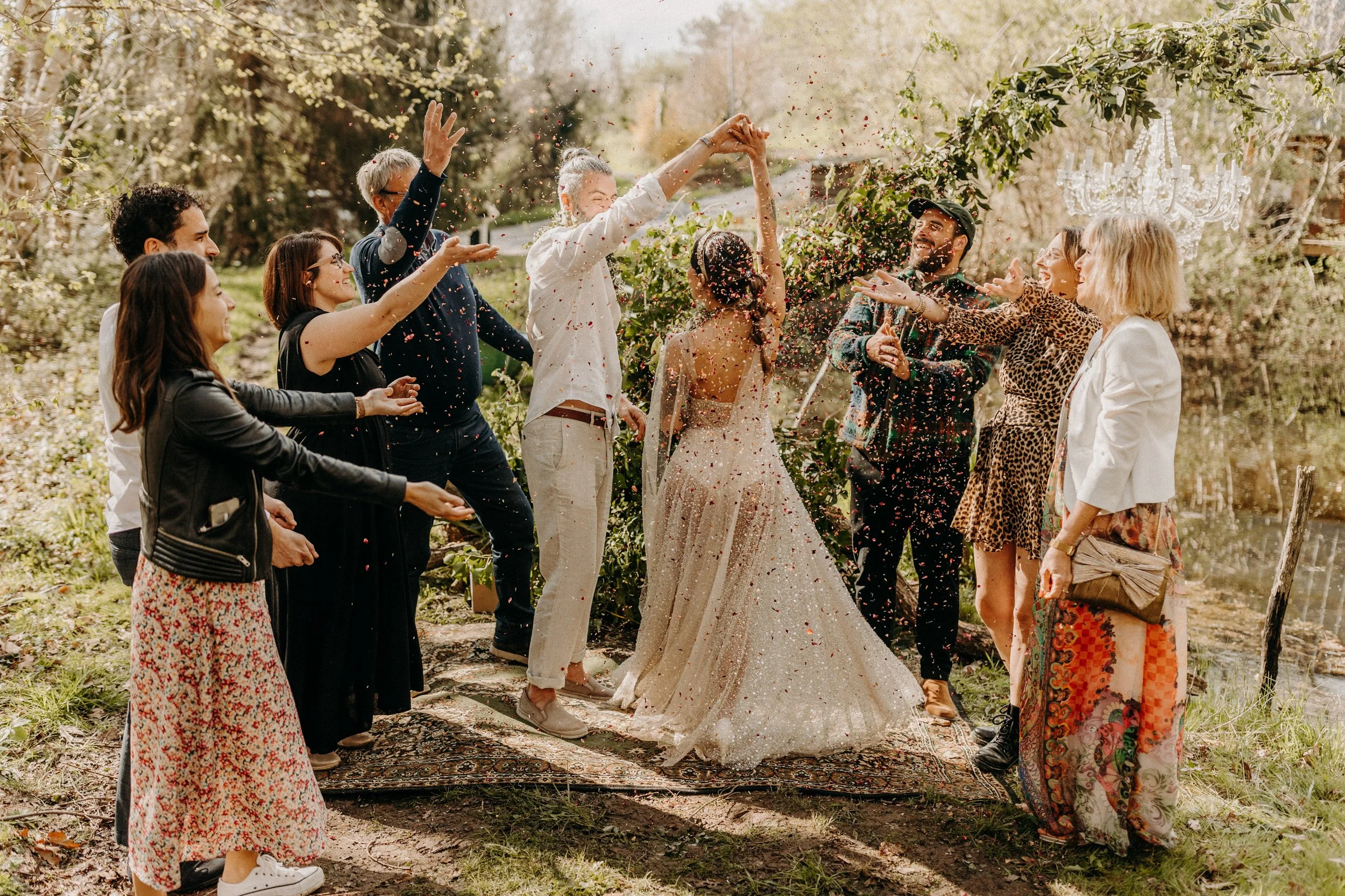 Un groupe de personnes fête un mariage en plein air, avec la mariée en robe de mariage et le marié entourés d'amis qui lancent des confettis.