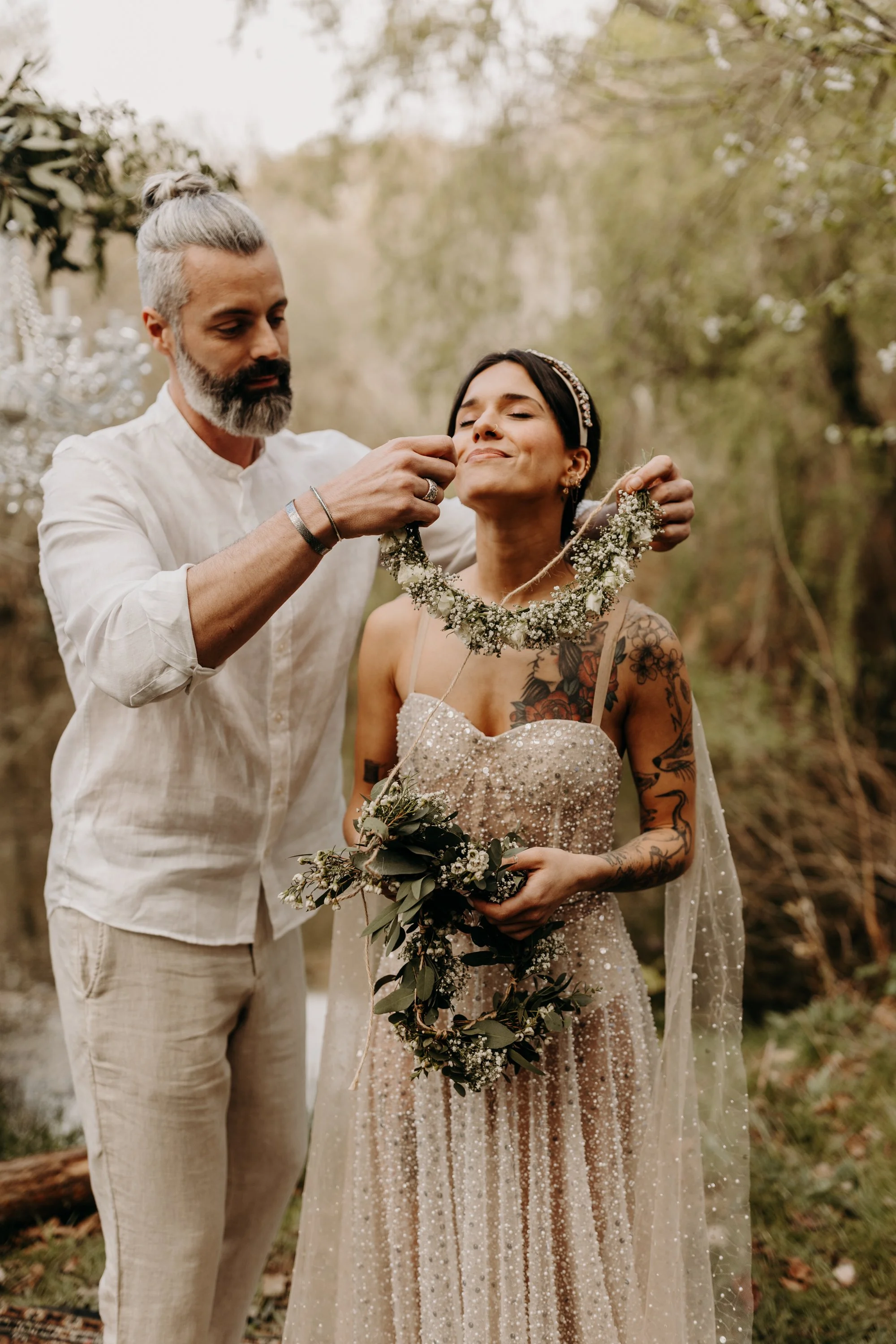 Un homme coiffé d'une coiffure grise et portant une chemise blanche aide une femme à mettre un collier de fleurs lors d'une cérémonie en plein air, avec un décor naturel en arrière-plan.