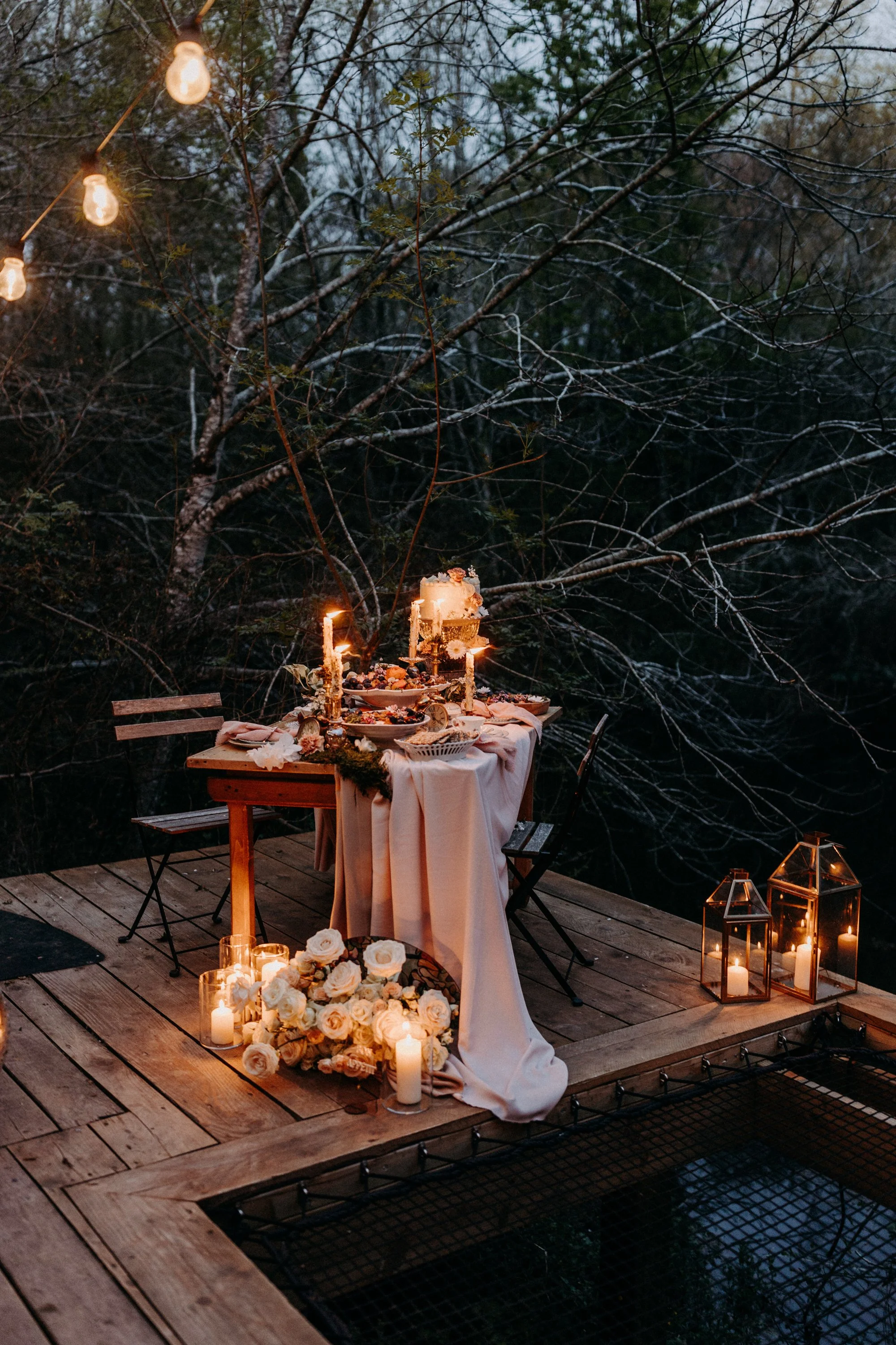 Une table dressée pour un repas romantique en plein air, décorée de bougies, de fleurs blanches, de chandeliers et d’un drapé rose pâle, entourée de lanternes en verre contenant des bougies, avec un fond de forêt en soirée.
