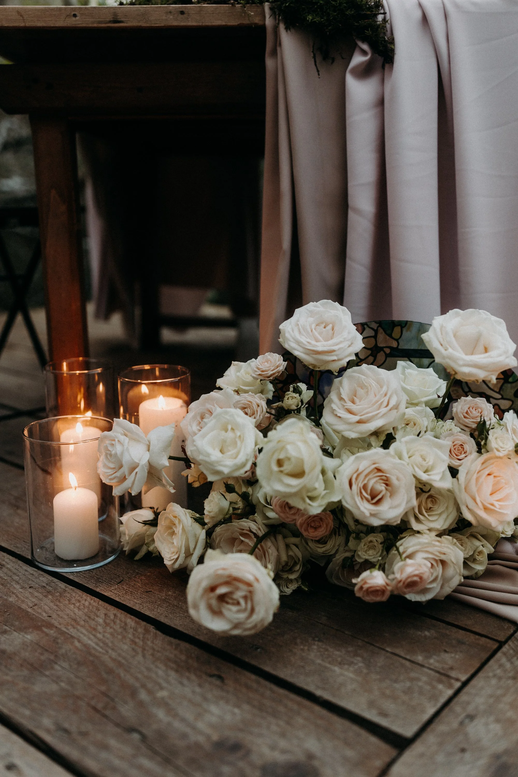 Un arrangement floral de roses blanches et roses pâles avec trois bougies pillées dans des votives en verre sur une table en bois