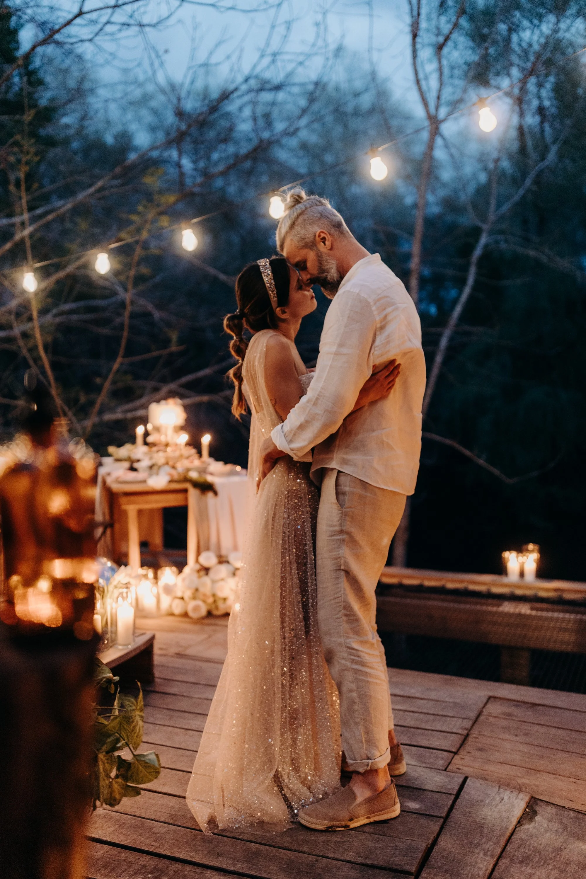 Un couple danse lors d'une cérémonie de mariage en plein air, entouré de lumières suspendues, avec une table de cérémonie décorée de fleurs et de bougies, en soirée.