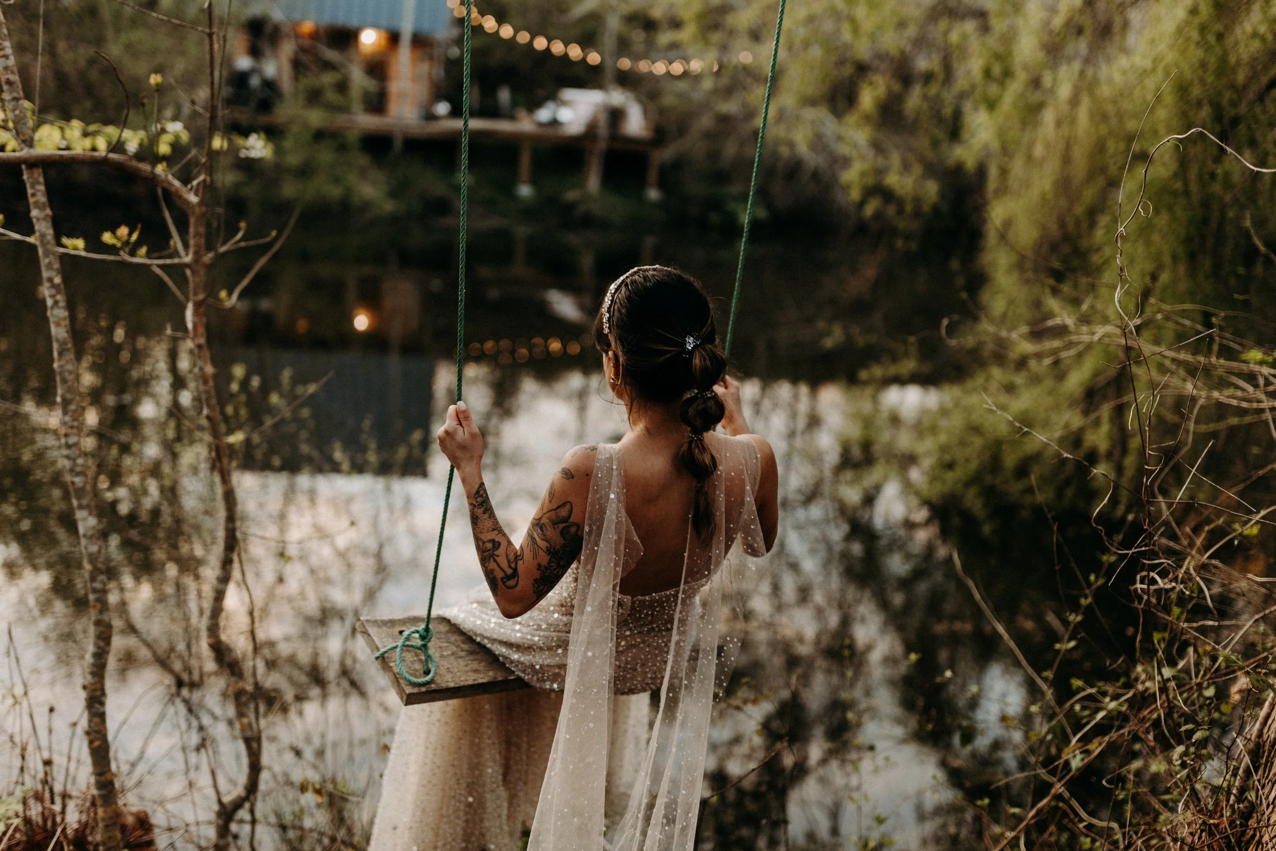 Une femme en robe de soirée assise sur une balançoire en bois au bord d'un lac, entourée de végétation, regarde au loin, scène en fin d'après-midi ou au coucher du soleil.